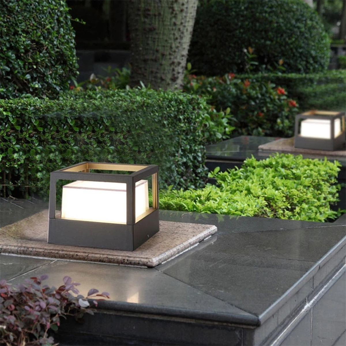 Garten mit zwei quadratischen Lampen auf Steinplattformen. Büsche und Bäume im Hintergrund. Dunkelgraue und grüne Farben.