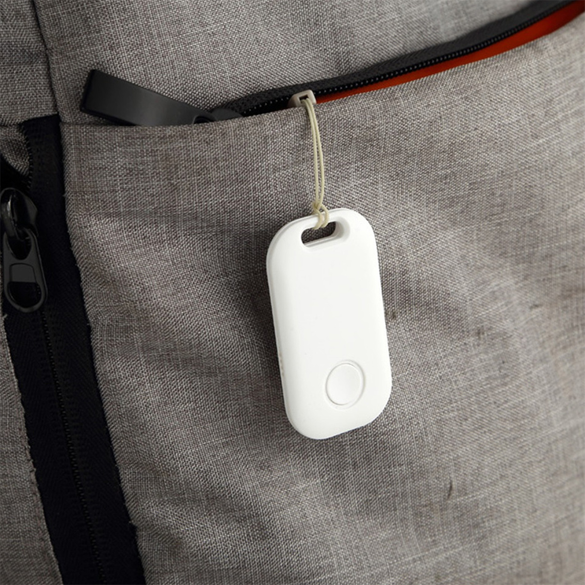 Ein weißer Tracker mit Loch und Knopf, an einer grauen Tasche befestigt, an einem grauen Rucksack.