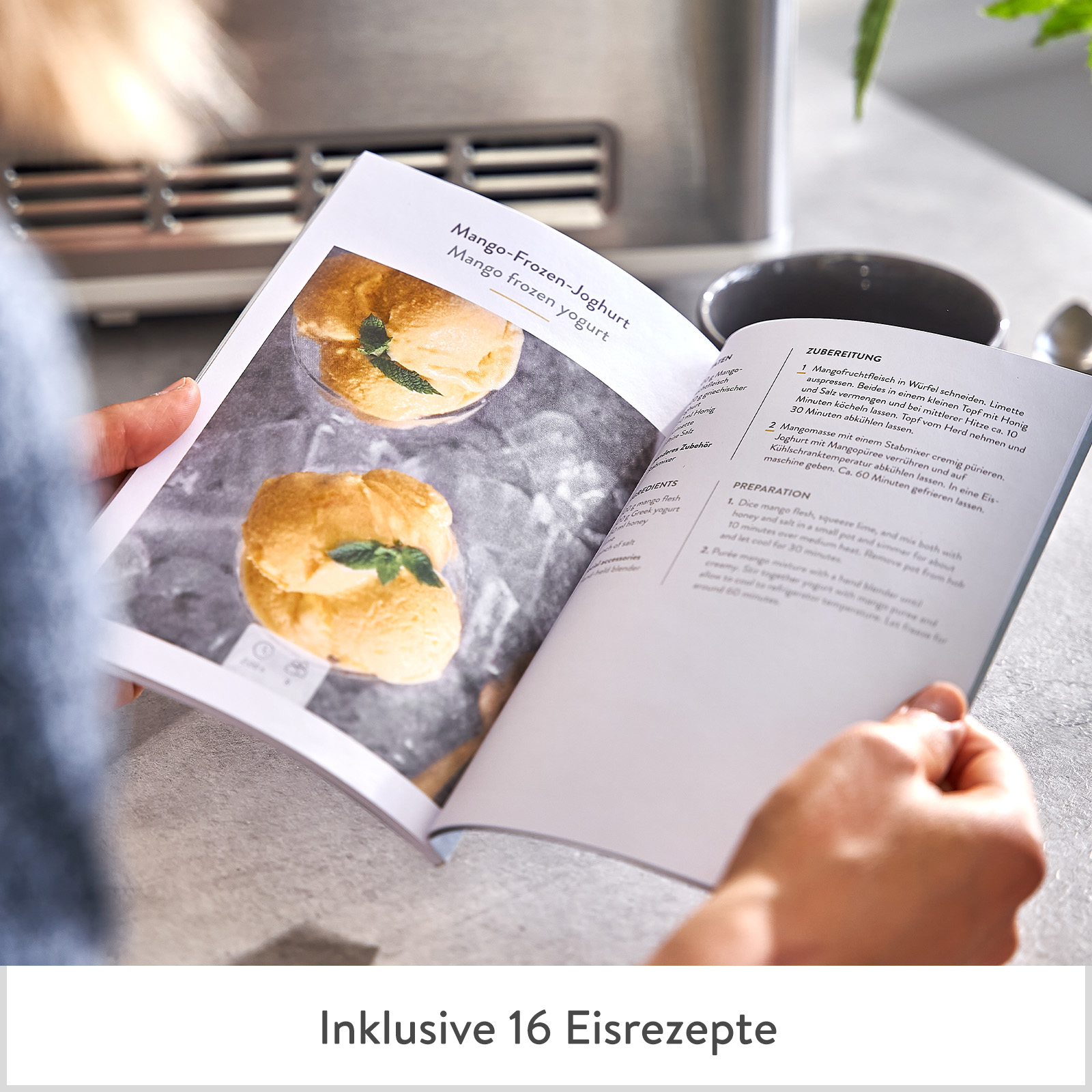 Offenes Kochbuch mit Mango-Frozen-Joghurt-Rezept. Hände halten es. Graue Oberfläche und eine Stahlarbeitsplatte.