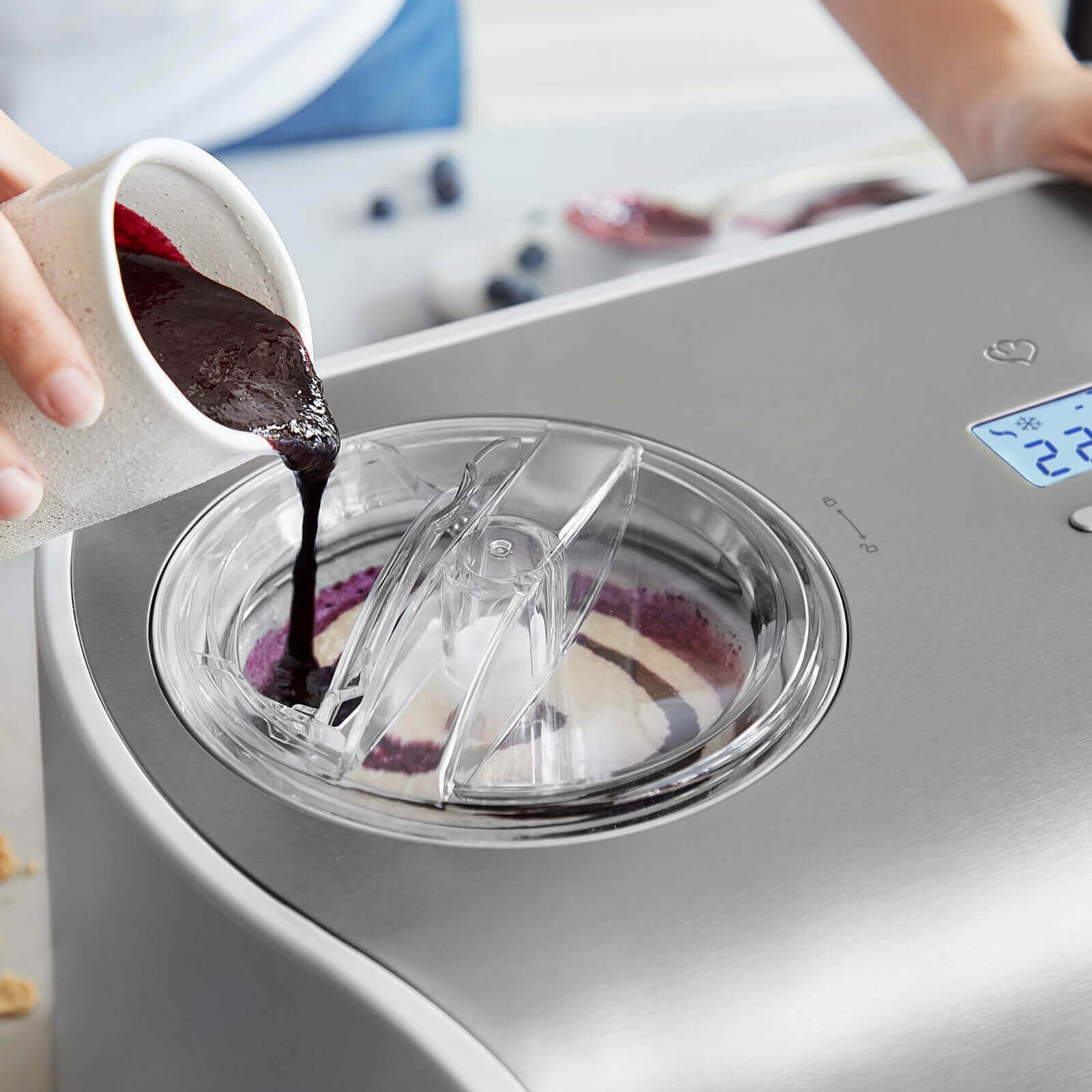 Eine Person gießt eine dunkle Flüssigkeit aus einem weißen Krug in eine Eismaschine. Die Maschine ist silber.
