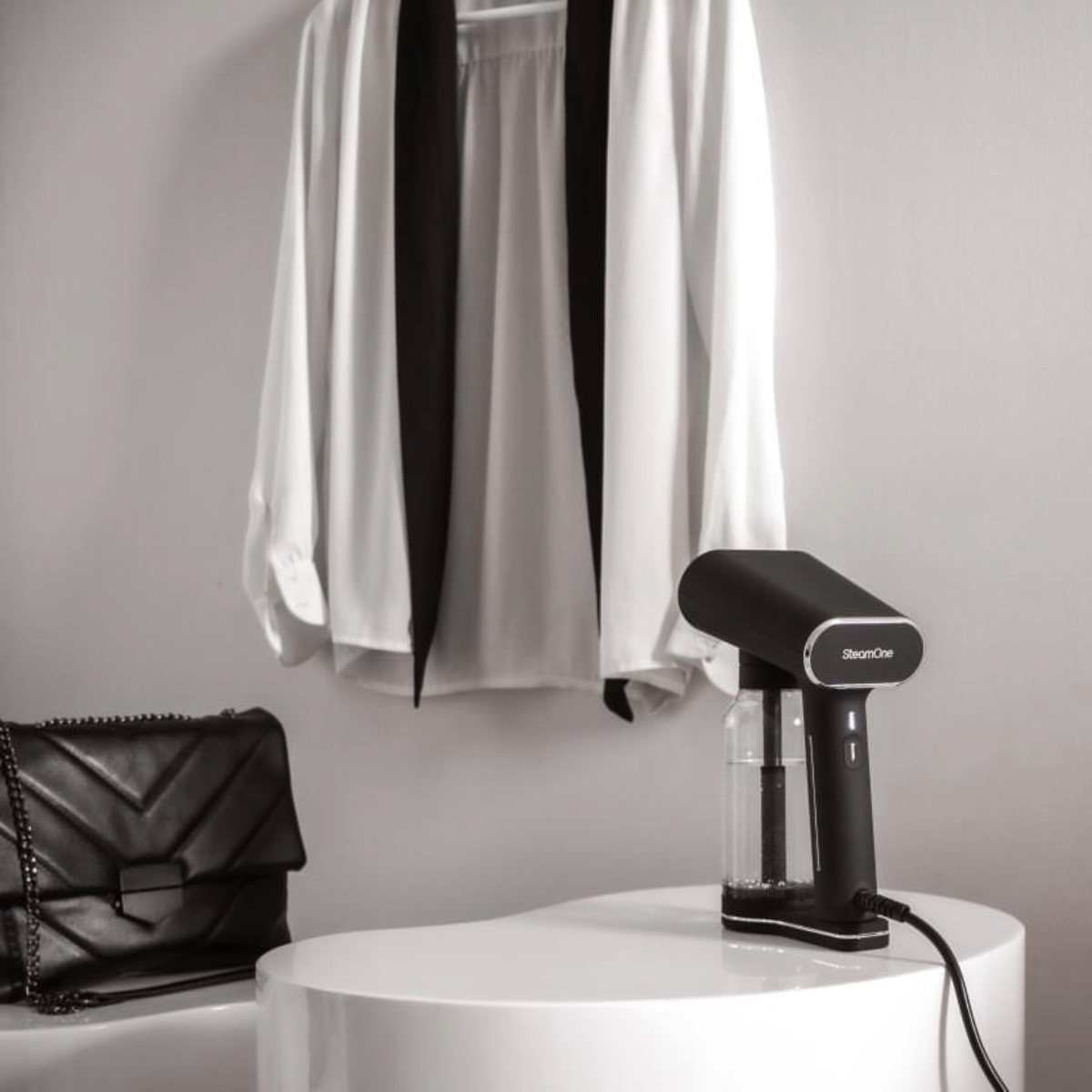 Image en noir et blanc d'un défroisseur noir à côté d'un sac à main et d'une chemise blanche sur un cintre.