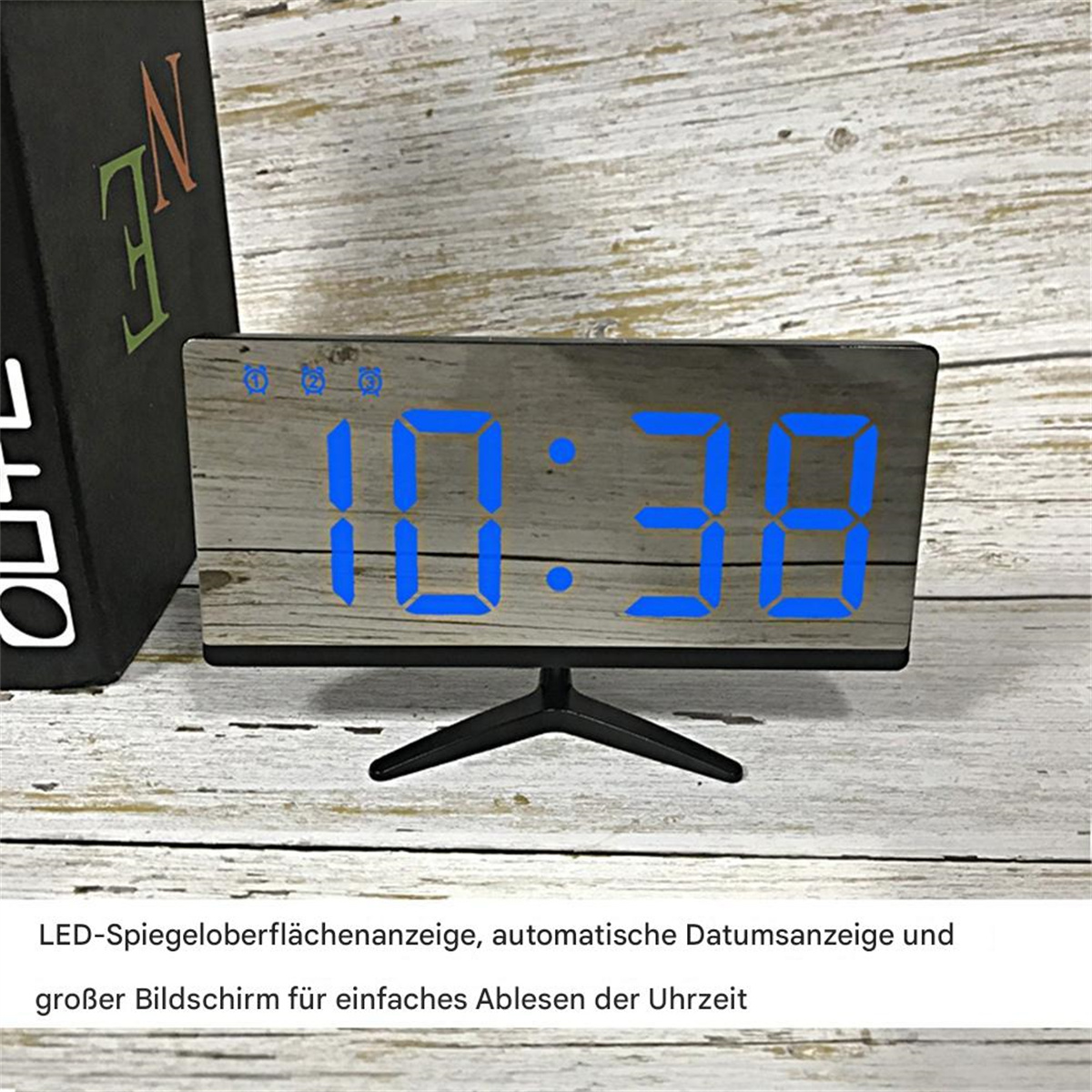 Eine Digitaluhr mit blauen Ziffern auf einer Holzoptik-Oberfläche. Sie zeigt 10:38 an. Im Hintergrund ist ein Buch.