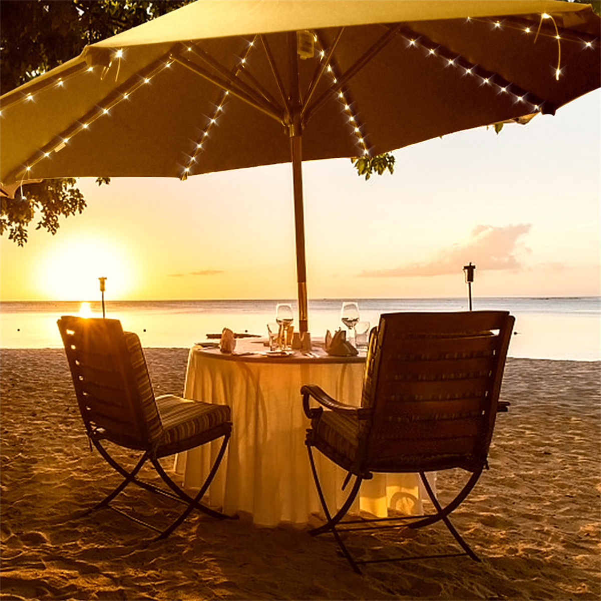 Strandszene bei Sonnenuntergang mit Tisch, Stühlen und Schirm mit Lichterketten.