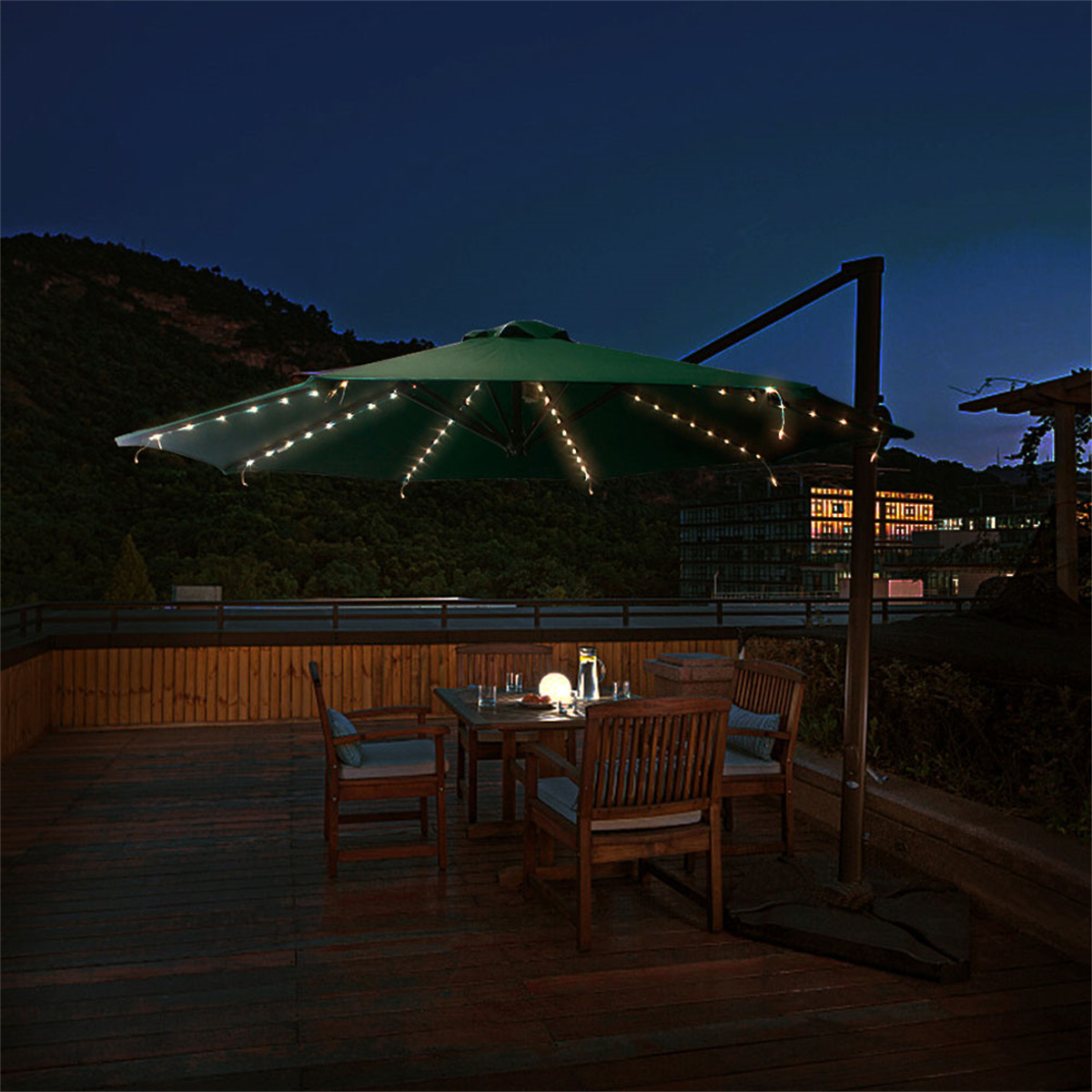 Außenszene bei Nacht mit beleuchtetem grünen Sonnenschirm, Tisch, Stühlen und Lichterketten auf der Terrasse.