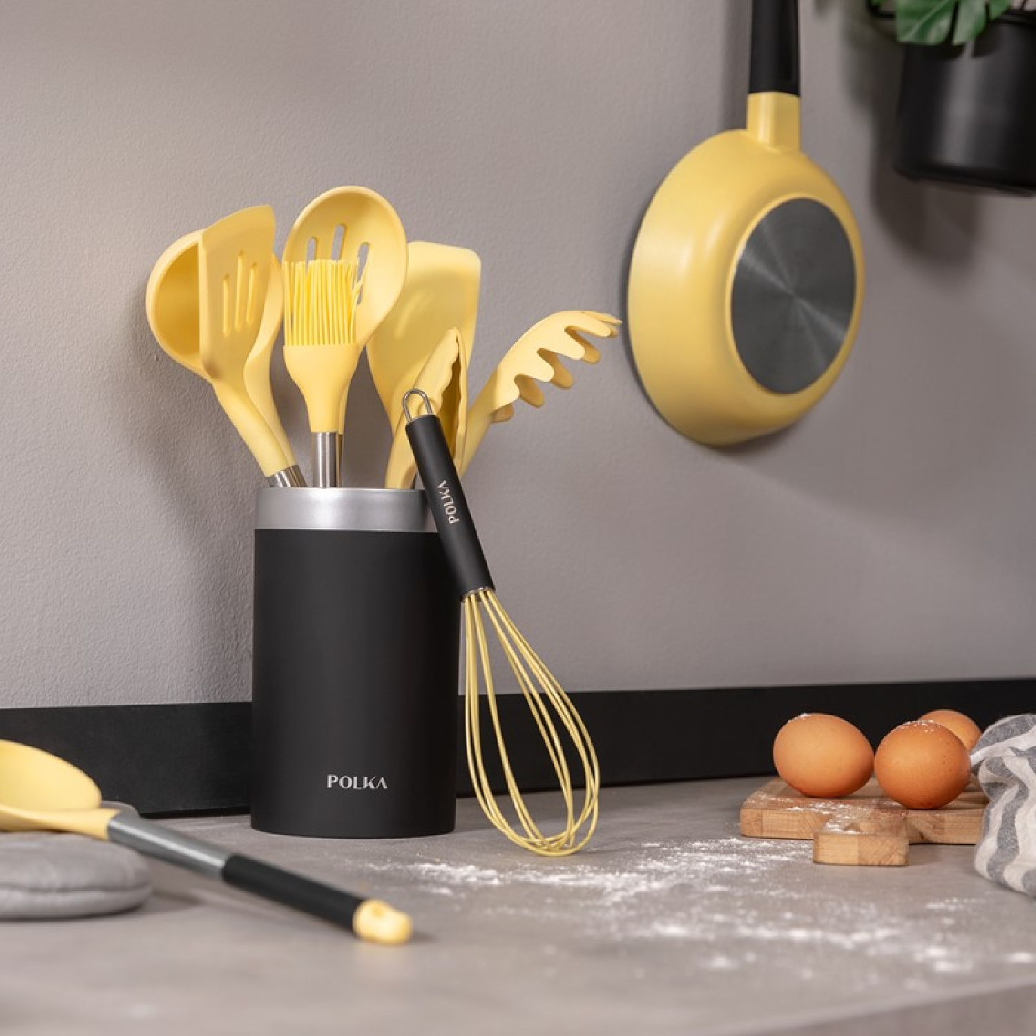 Utensilios de cocina amarillos en un soporte negro, con huevos y sartén en la encimera.