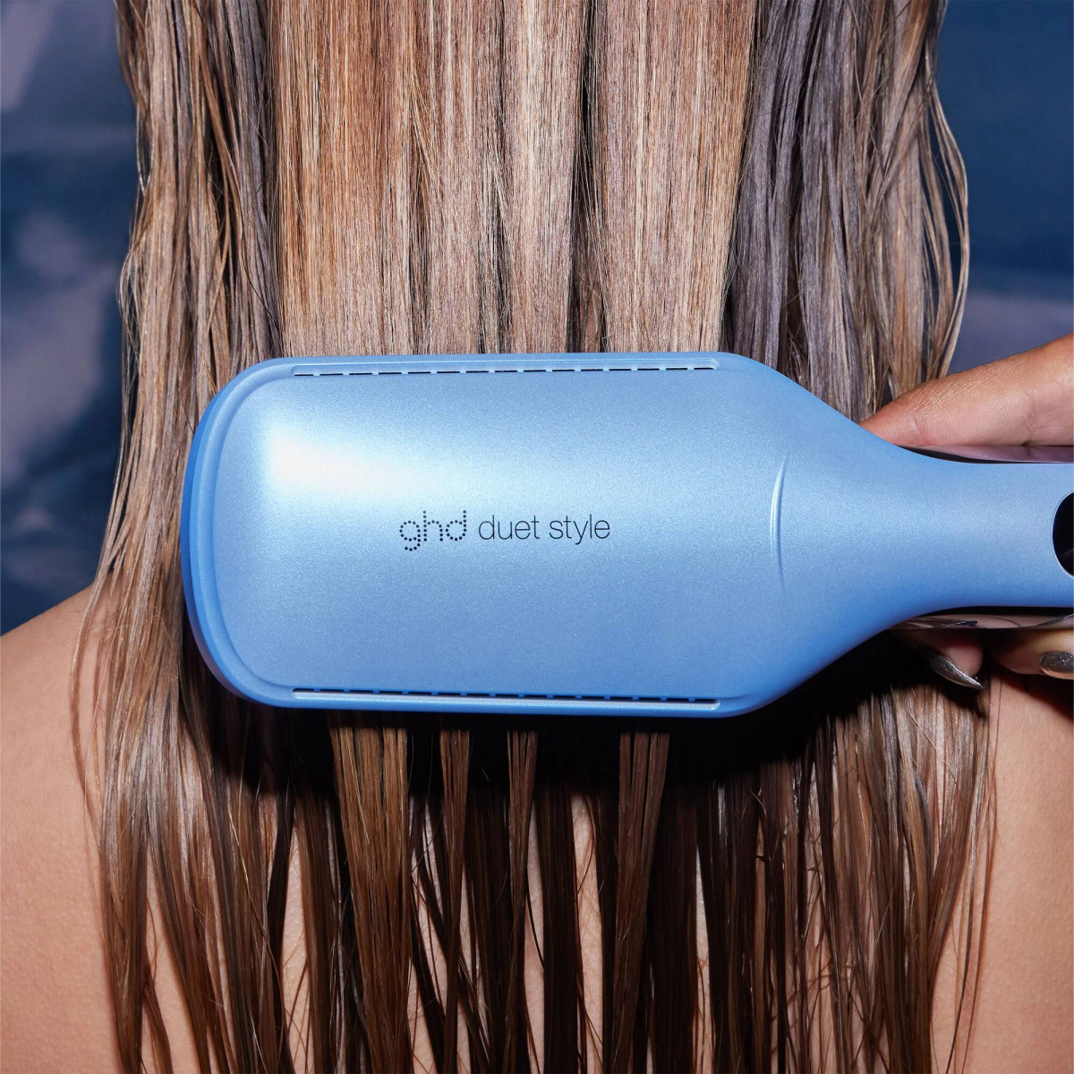 Una mano usa una piastra ghd duet style blu su capelli bagnati.