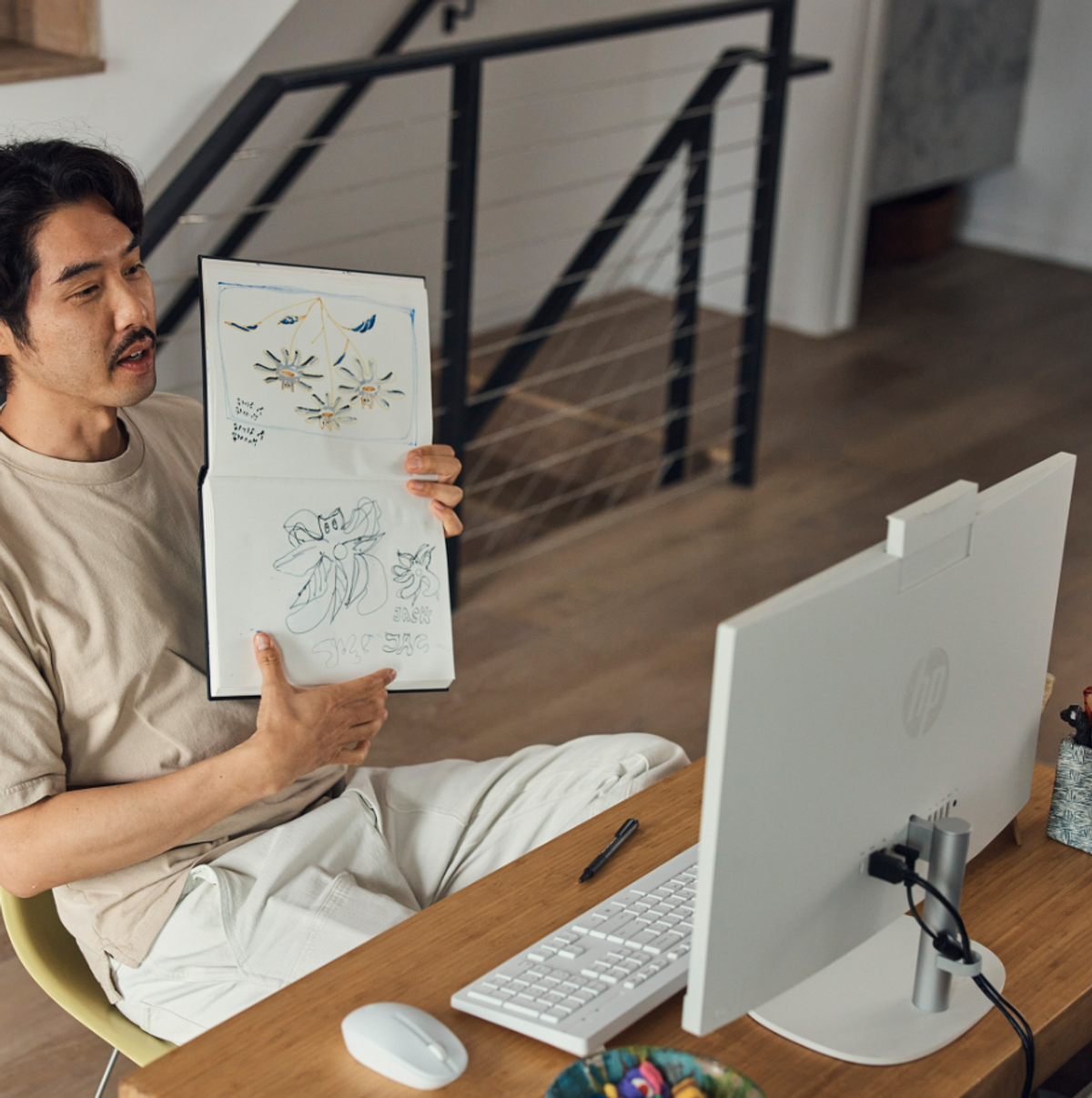 Hombre mostrando dibujos en una libreta durante una videollamada. Un monitor y teclado blancos están en el escritorio.