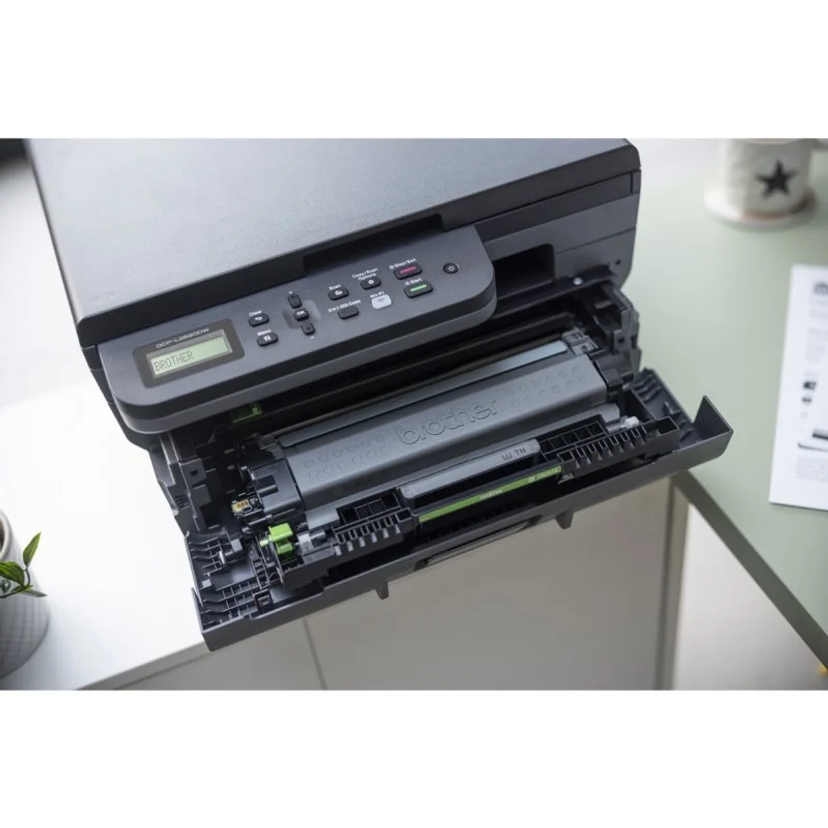 Drukarka Brother z otwartą szufladą. W środku czarny wkład z tonerem. Widoczny biały stół.