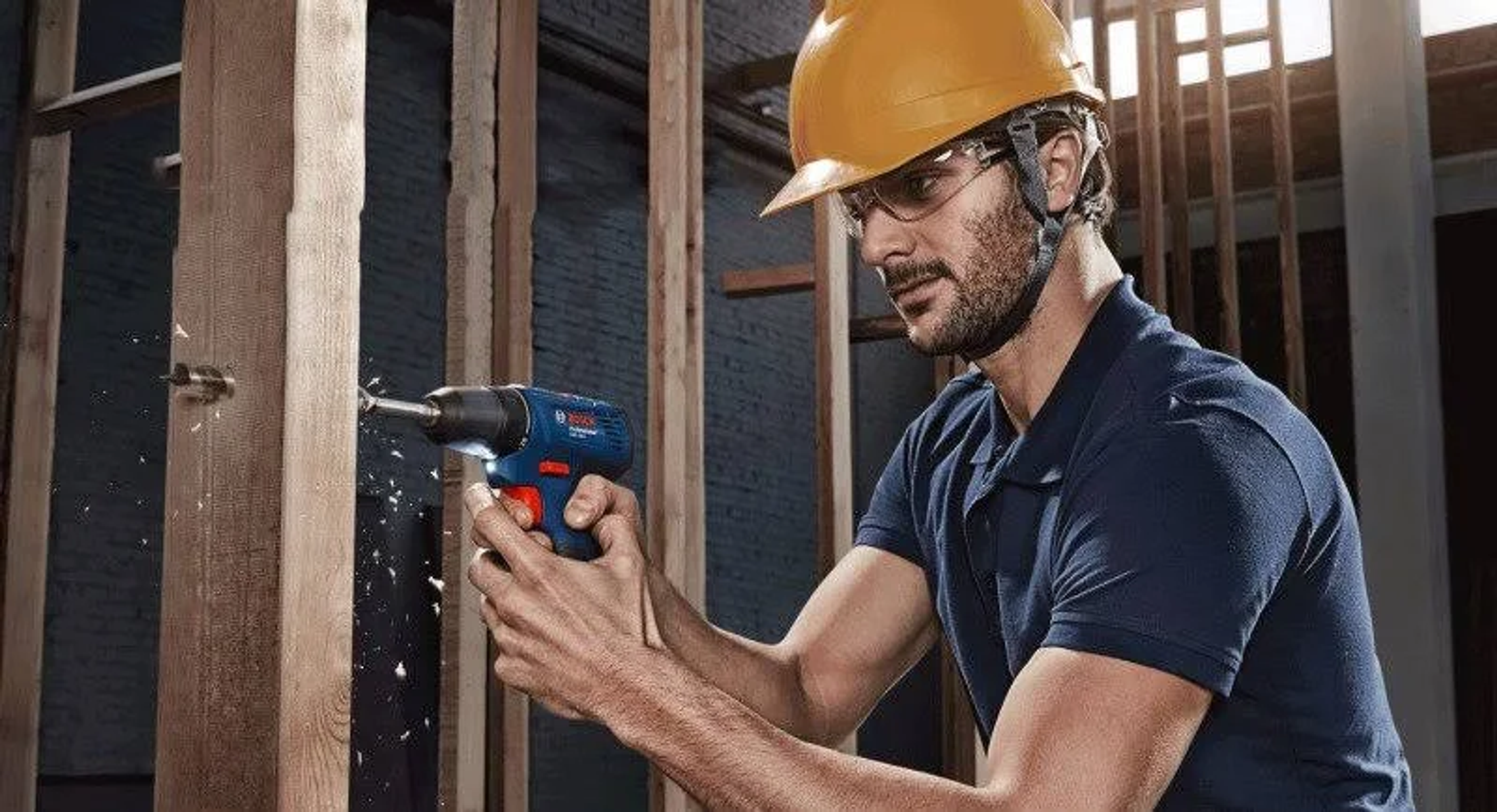 Un carpentiere fora il legno con un trapano blu e rosso, indossando un casco.