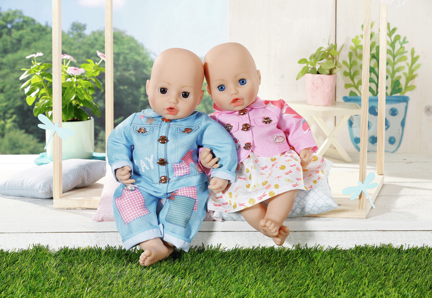 Zwei Babypuppen, eine in Blau, eine in Rosa, sitzen auf einer Schaukel. Grünes Gras im Hintergrund.