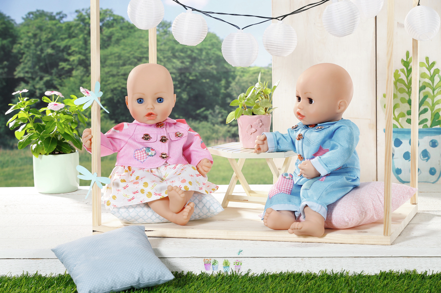 Zwei Babypuppen in einem Holzhaus. Eines trägt Rosa, das andere Blau, auf Gras mit weißen Laternen.