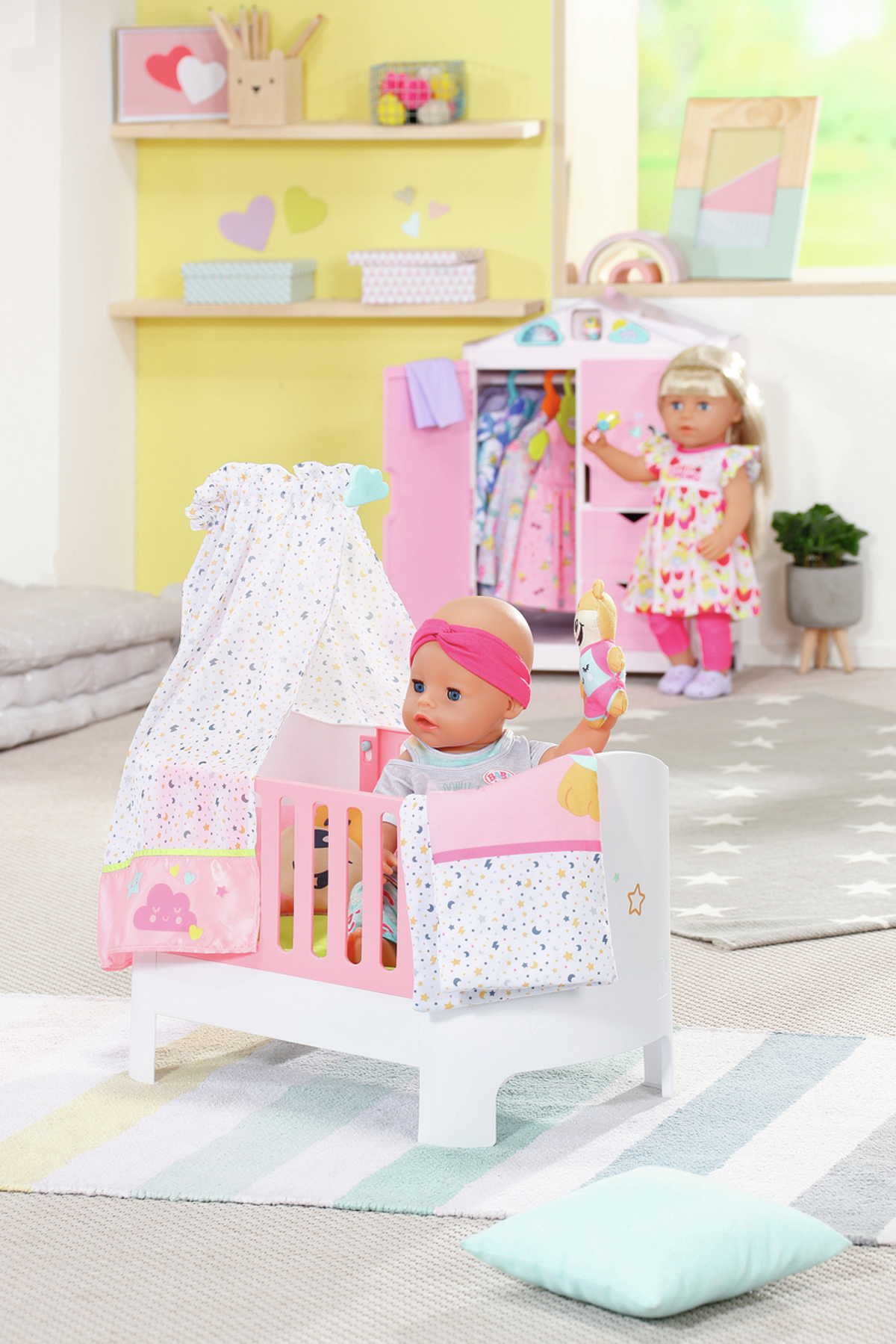 Babypuppe im Kinderbett mit Baldachin. Eine andere Puppe steht in der Nähe. Ein rosa Schrank und Regale sind im Hintergrund.