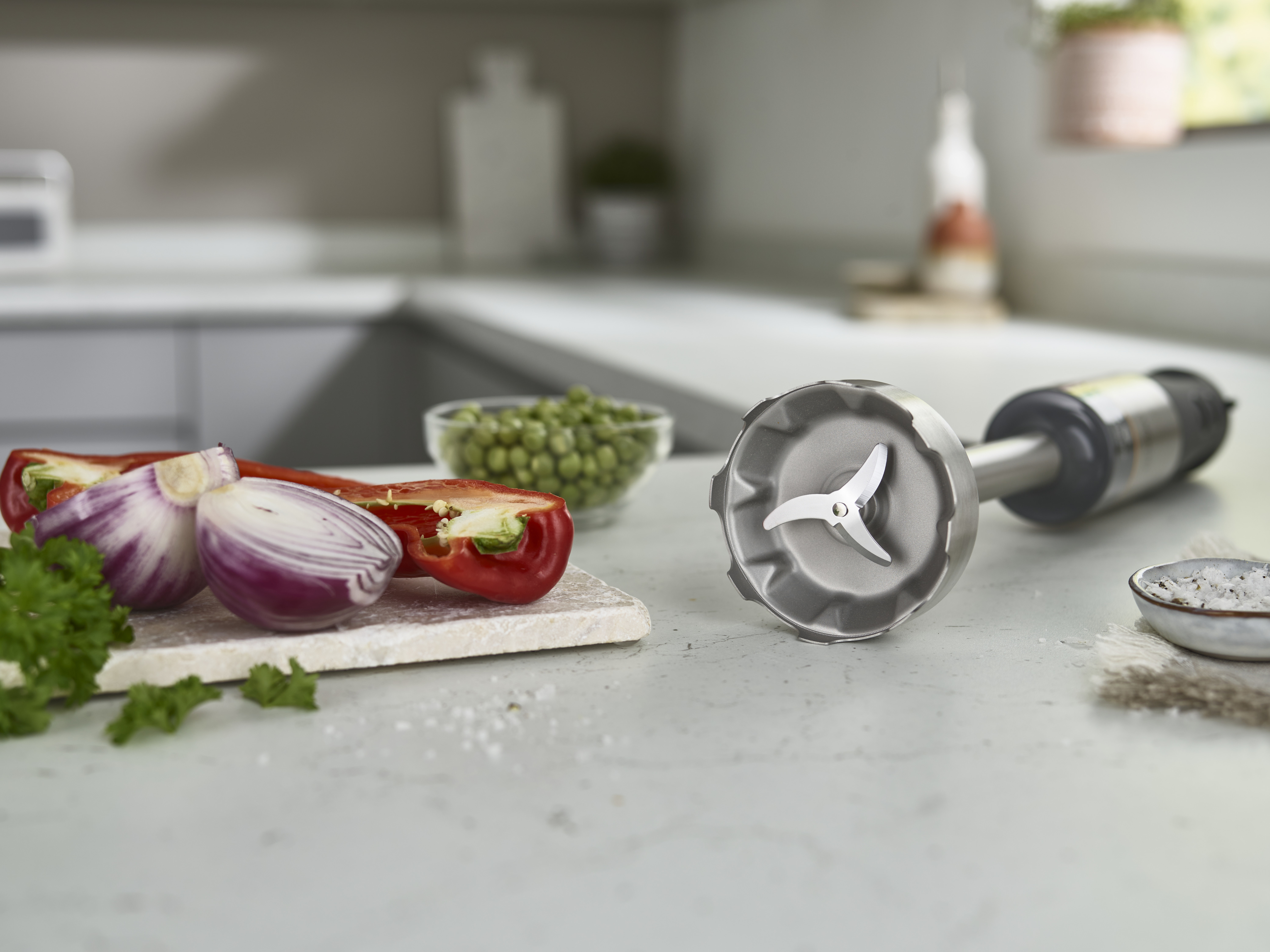 Stabmixer mit Gemüse auf einer Arbeitsplatte. Rote Zwiebeln, roter Paprika, Erbsen in einer Schüssel.