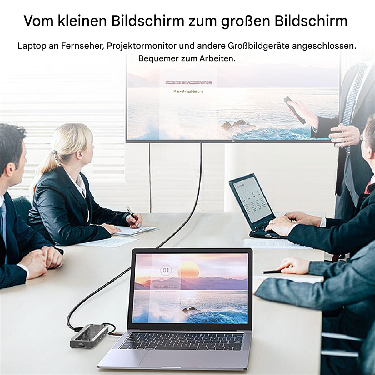Leute sitzen um einen Tisch und schauen auf einen Laptop-Bildschirm, der an einen Monitor angeschlossen ist. Der Laptop ist geöffnet.