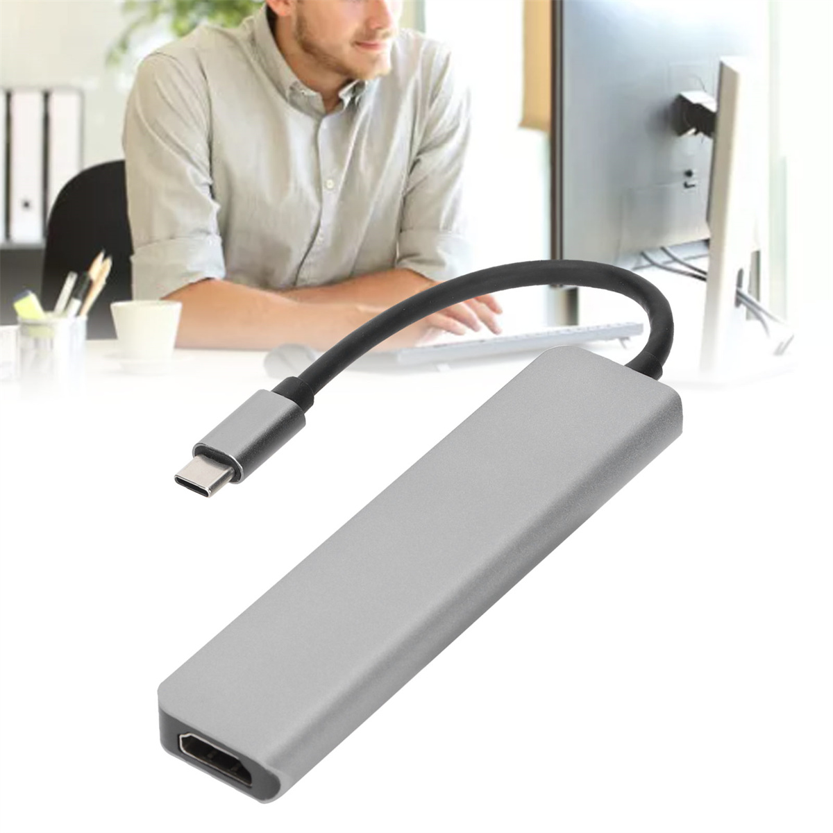 Ein silberner USB-C-Hub mit einem schwarzen Kabel, vor einem Mann und einem Monitor. Der Hub ist silber, mit USB-C-Anschluss.