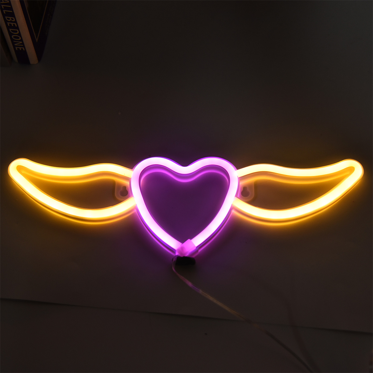 Neonherz mit Flügeln. Das Herz ist lila, die Flügel gelb, an einer dunklen Wand.