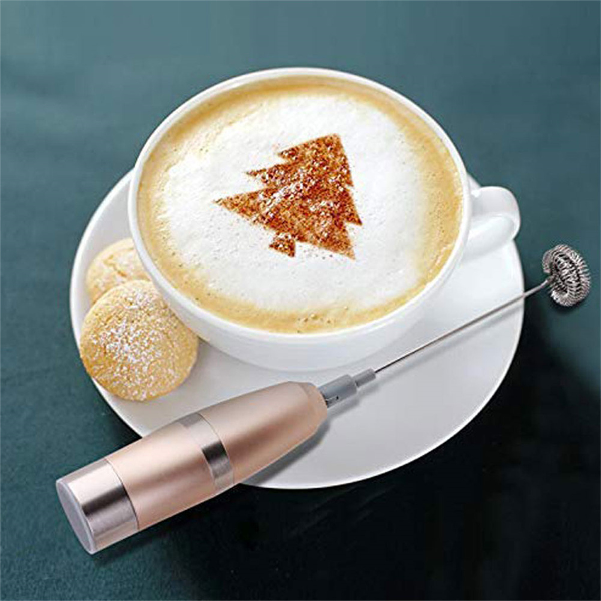 Eine Tasse Cappuccino mit Weihnachtsbaum-Design, serviert mit einem Milchaufschäumer und Keksen.