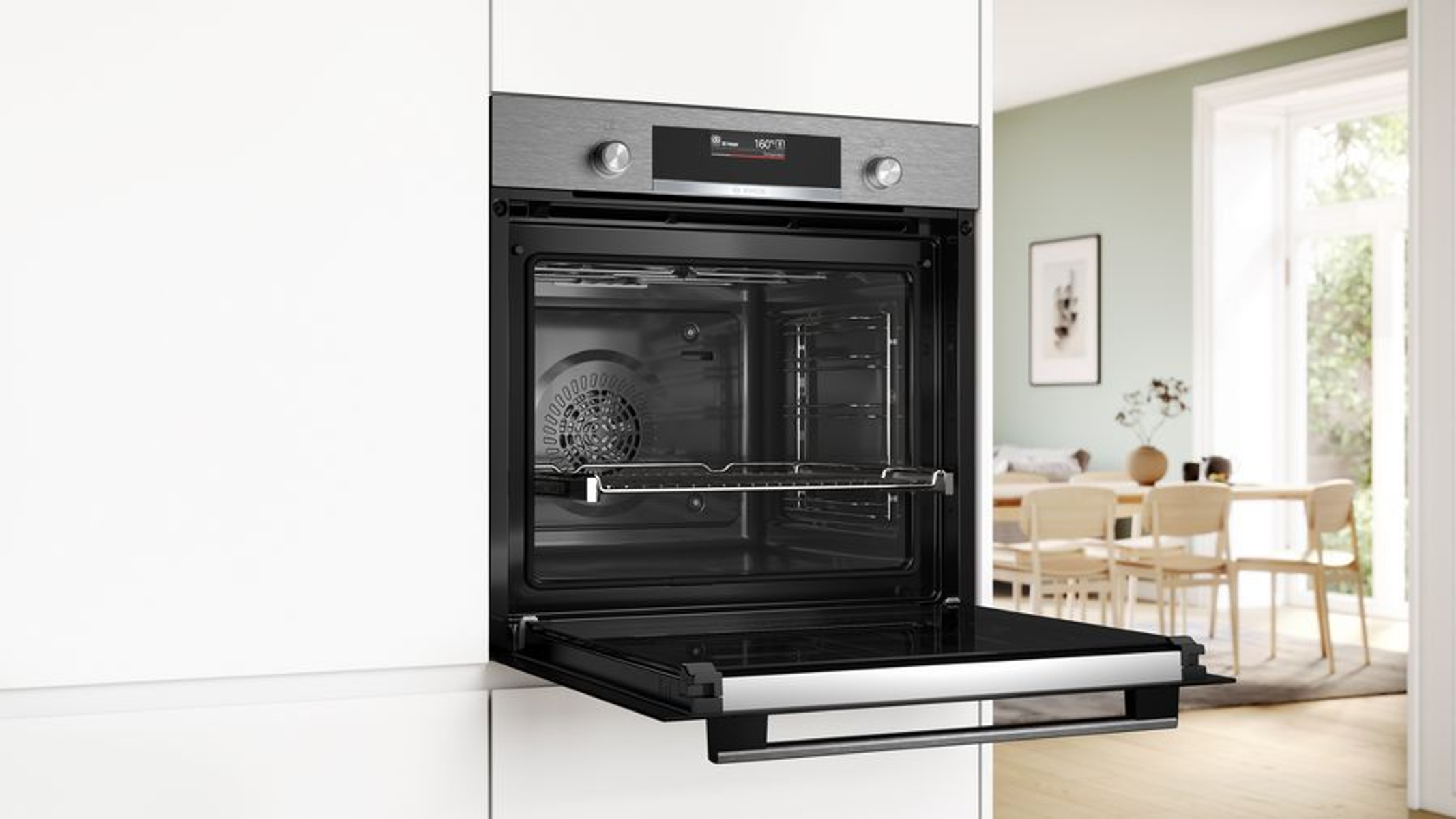 Horno empotrado con puerta abierta, mostrando interior con rejilla. Armarios blancos y comedor visibles.