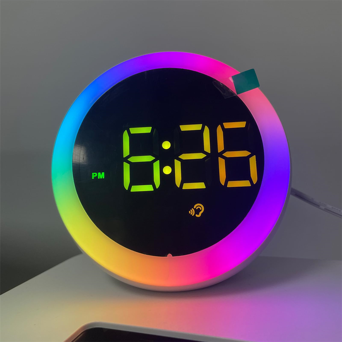 Digitaler Wecker mit 6:26 PM. Regenbogenfarbiger Lichtring um den schwarzen Bildschirm. Weiße Oberfläche.