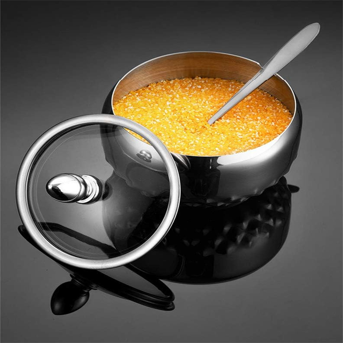 Eine silberne Schüssel mit einem durchsichtigen Deckel und einem Löffel. Die Schüssel enthält gelbe Granulate. Dunkler Hintergrund.