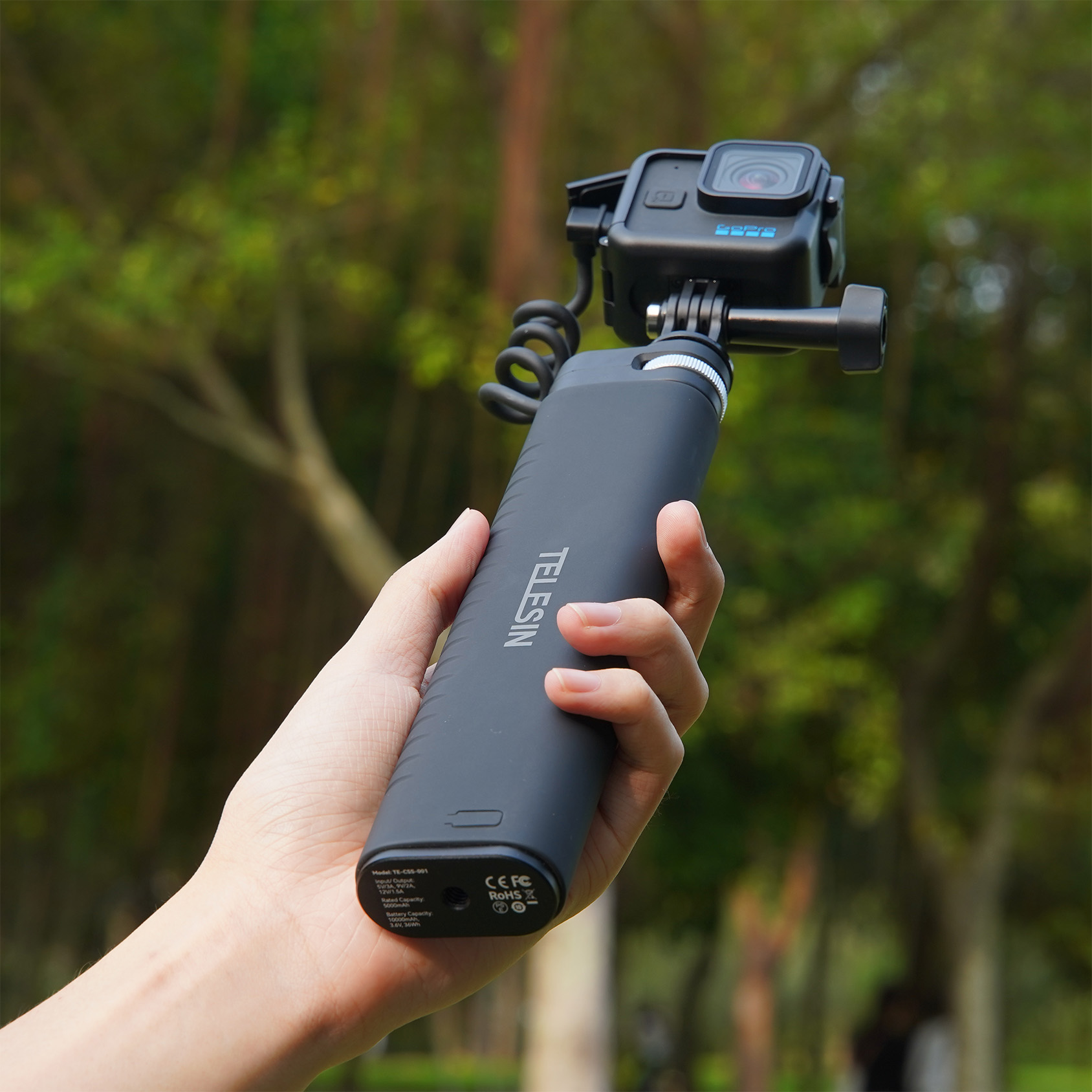Een persoon die een selfiestick vasthoudt met een camera eraan, in een park.