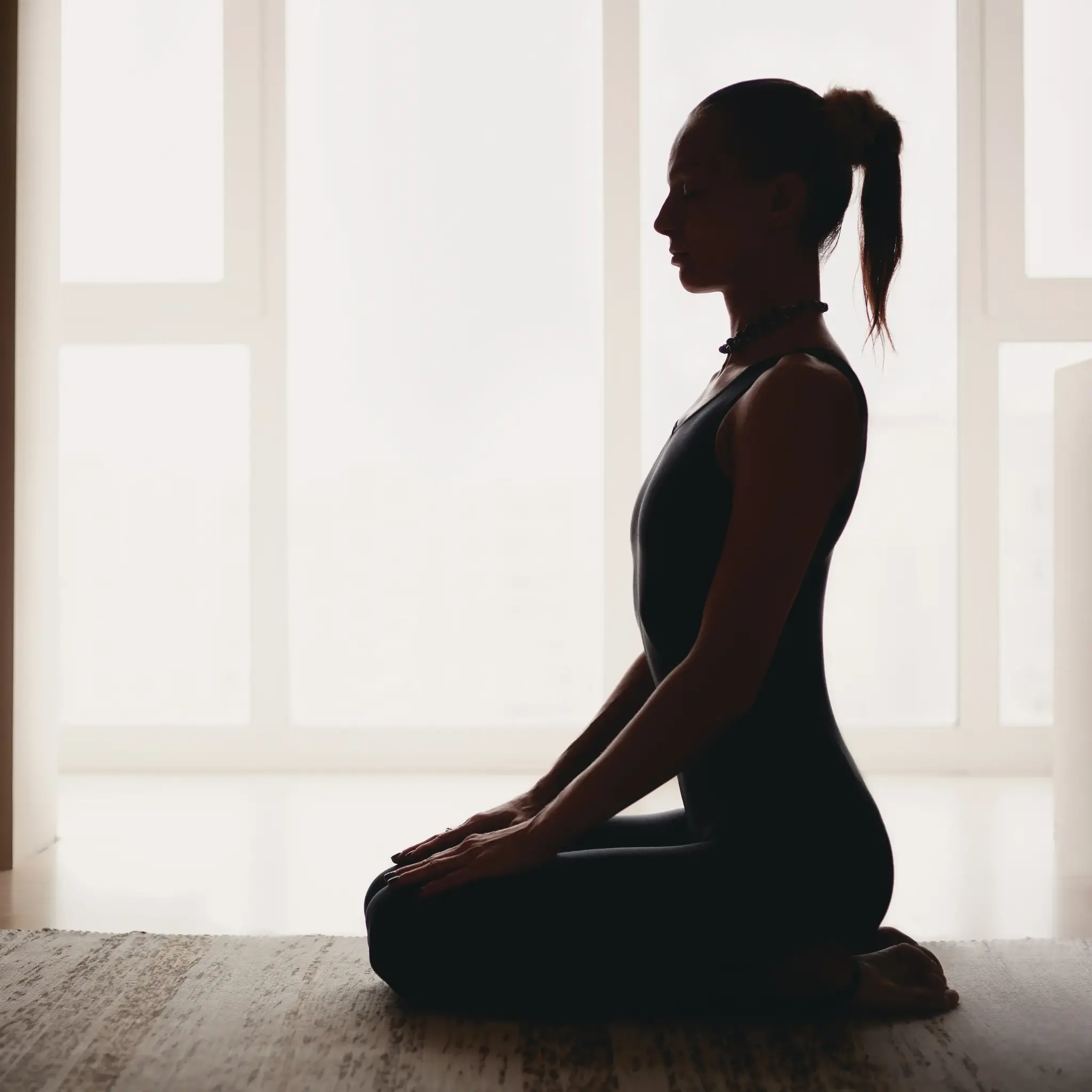 Silhouette of a woman in a yoga pose. She is kneeling in front of a bright window.
