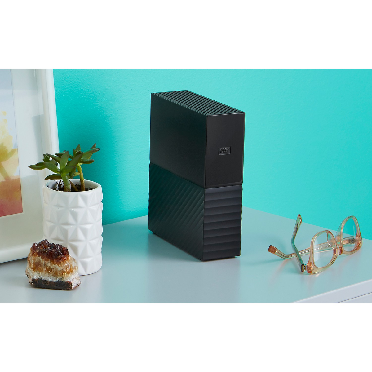 Czarny zewnętrzny dysk twardy na szarej powierzchni, z białą ramą, rośliną i okularami w tle.