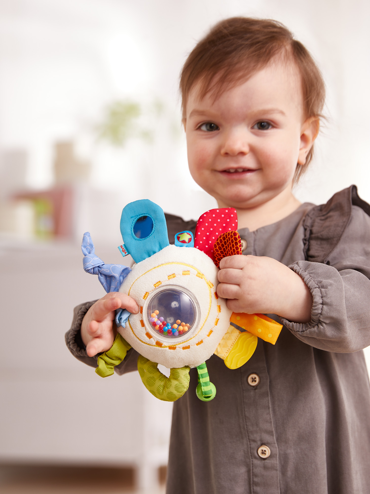 Ein lächelndes Baby hält ein farbenfrohes Sensorikspielzeug mit Perlen und Texturen.