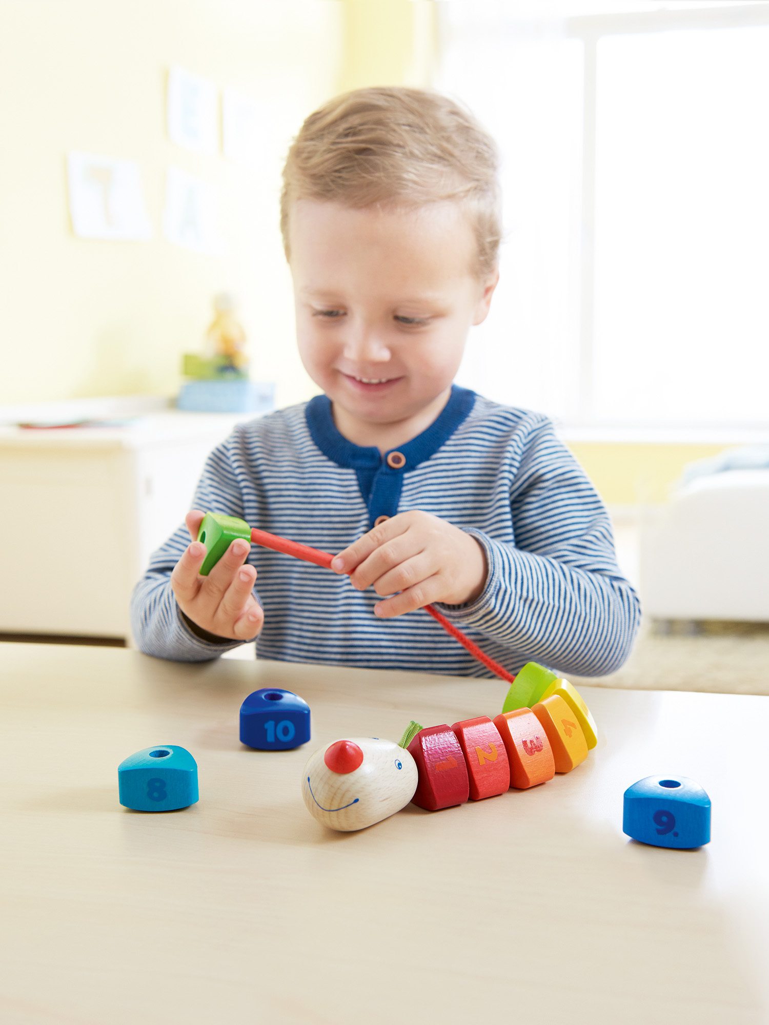 Ein Junge spielt an einem Tisch mit einem Holzraupenspielzeug. Er lächelt und hält einen grünen Block.
