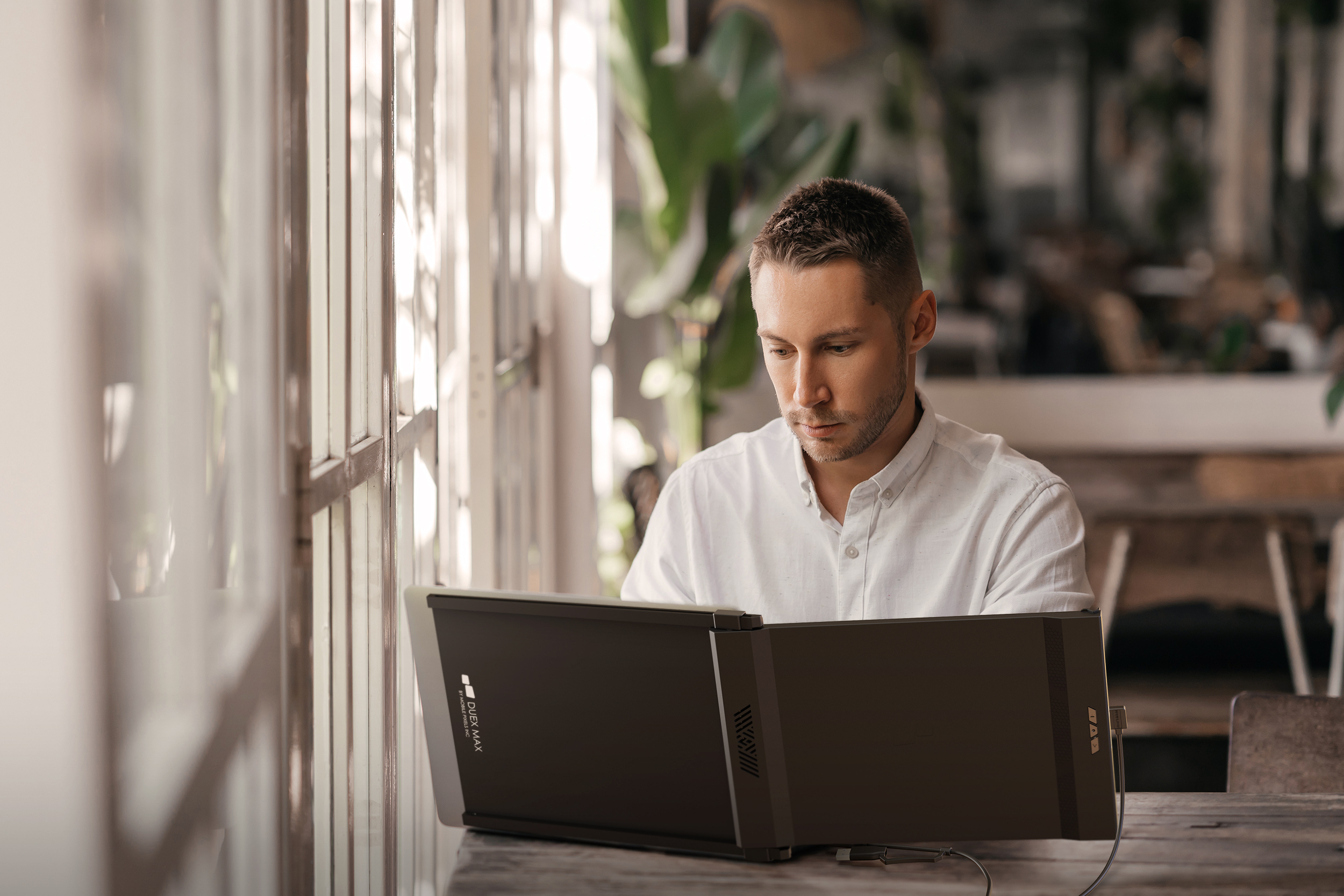 Mężczyzna korzysta z monitora z dwoma ekranami. Siedzi przy stole blisko okna. Światło jest dobre.