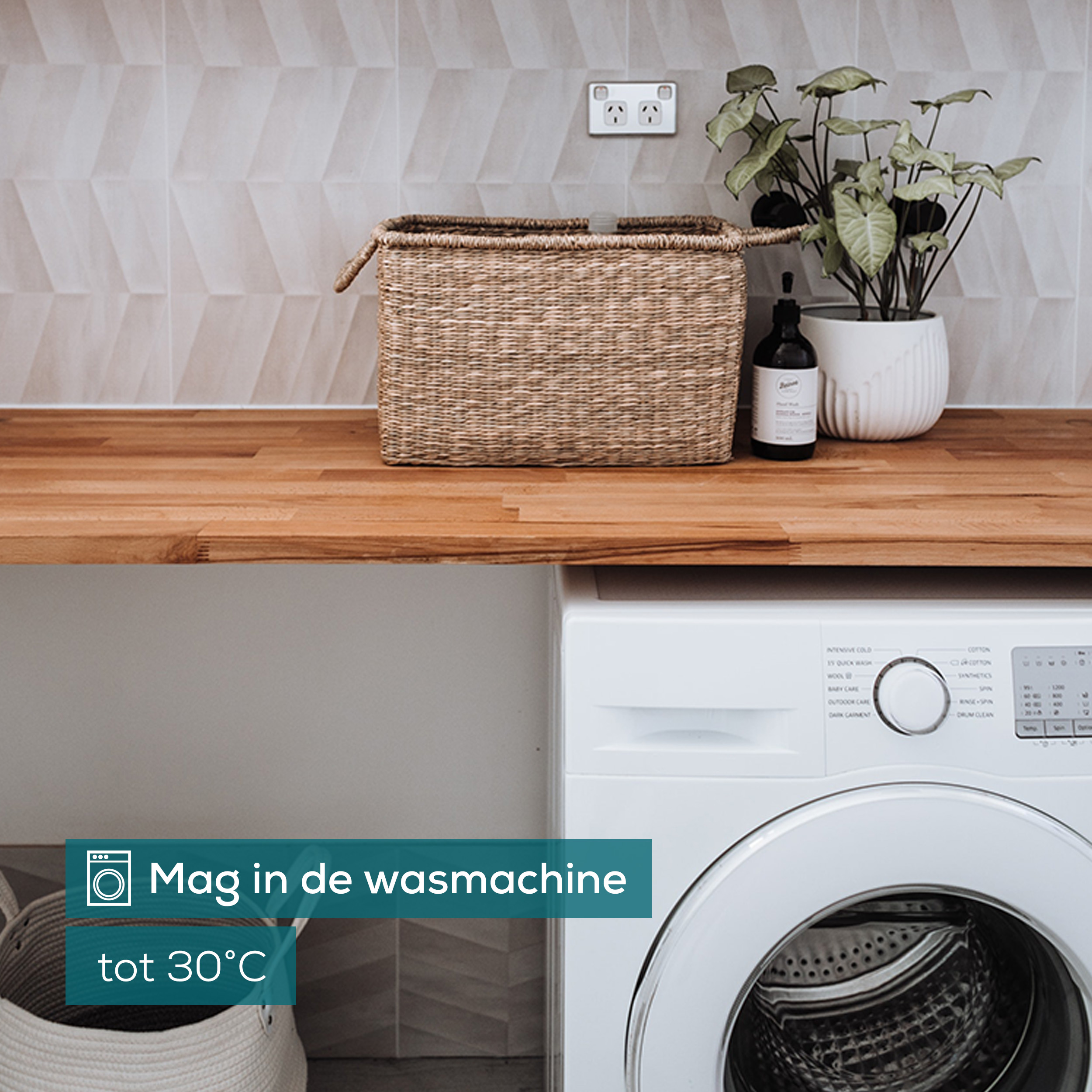 Scena di cucina con lavatrice. Sono visibili un cesto, una pianta e un piano di lavoro in legno. Testo: 'Mag in de wasmachine'.