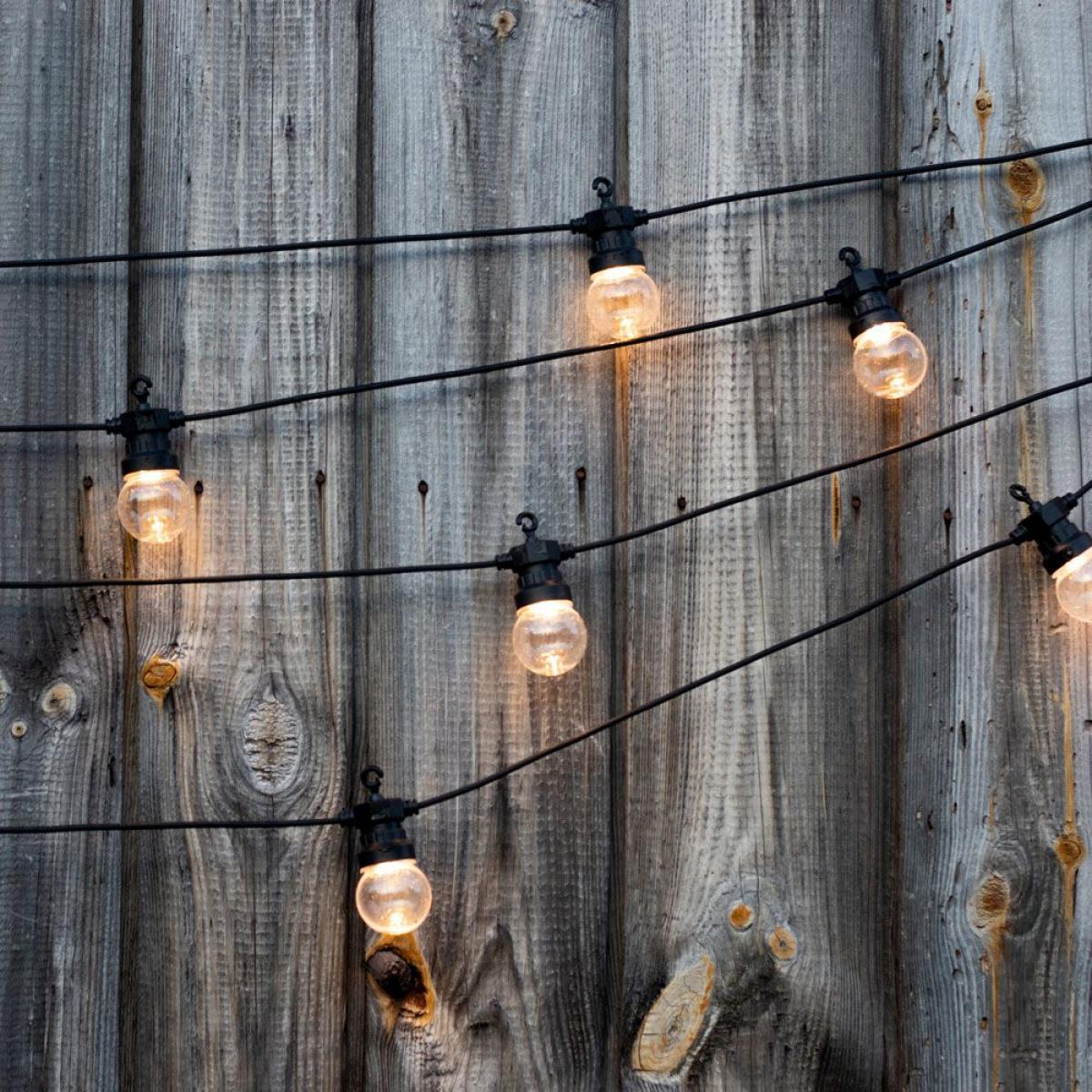 Catena luminosa con lampadine trasparenti su fili neri, contro una staccionata di legno stagionato. La luce è bianco caldo.
