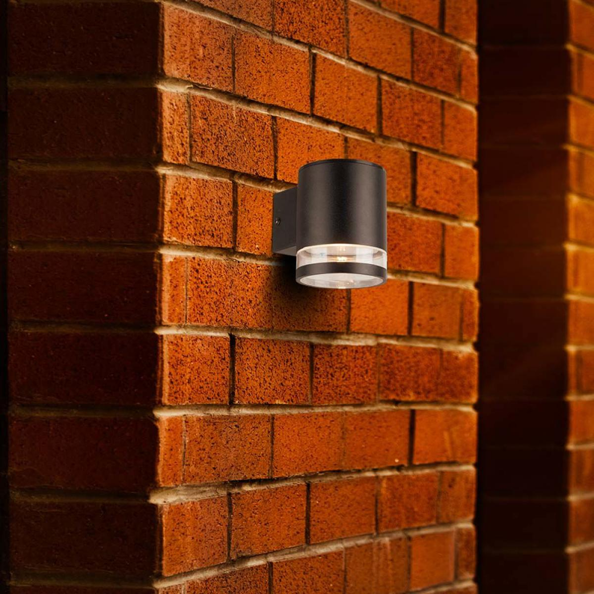 Eine schwarze zylindrische Wandleuchte an einer roten Backsteinmauer mit Licht.
