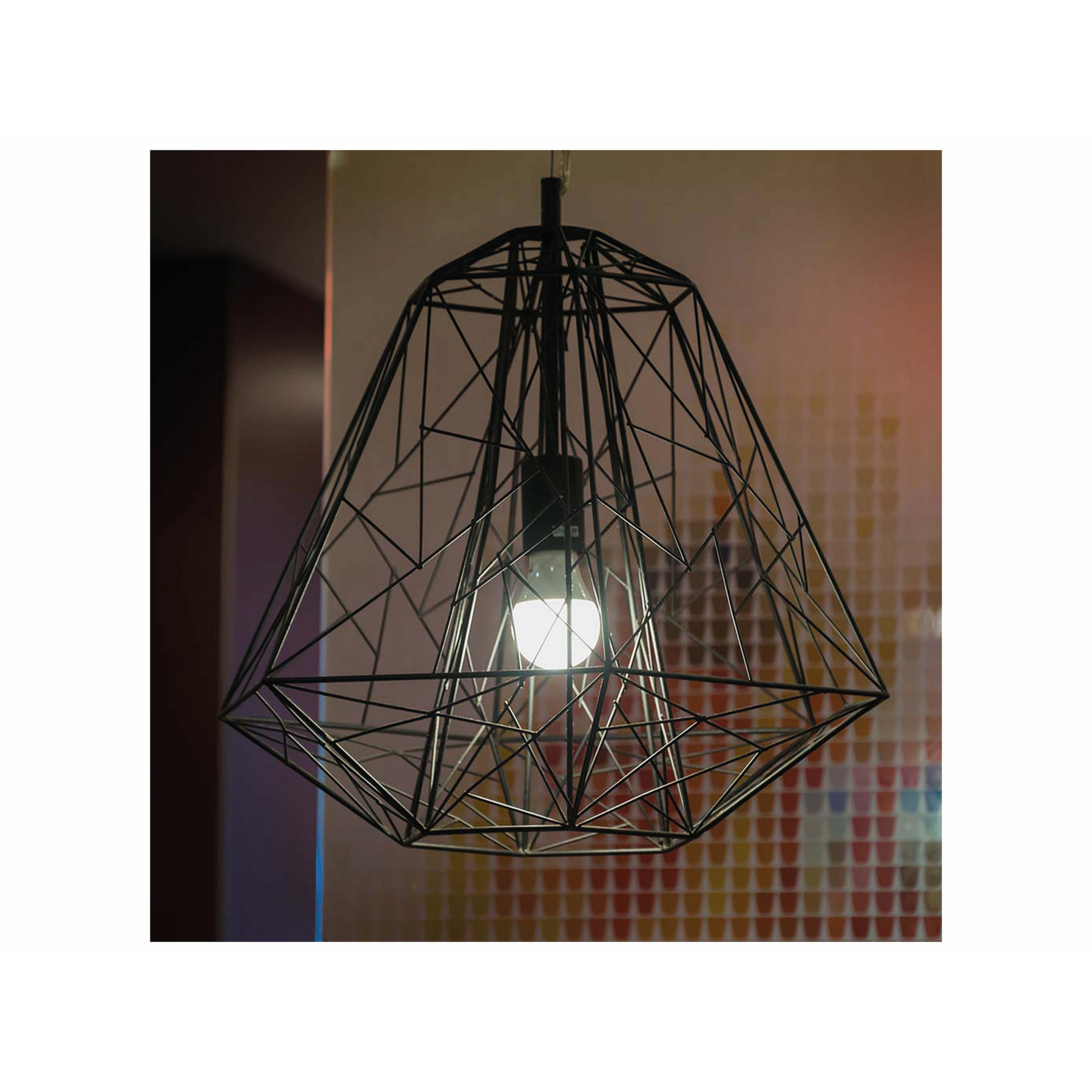 Un lampadario geometrico nero con una lampadina accesa all'interno. Lo sfondo è sfocato.