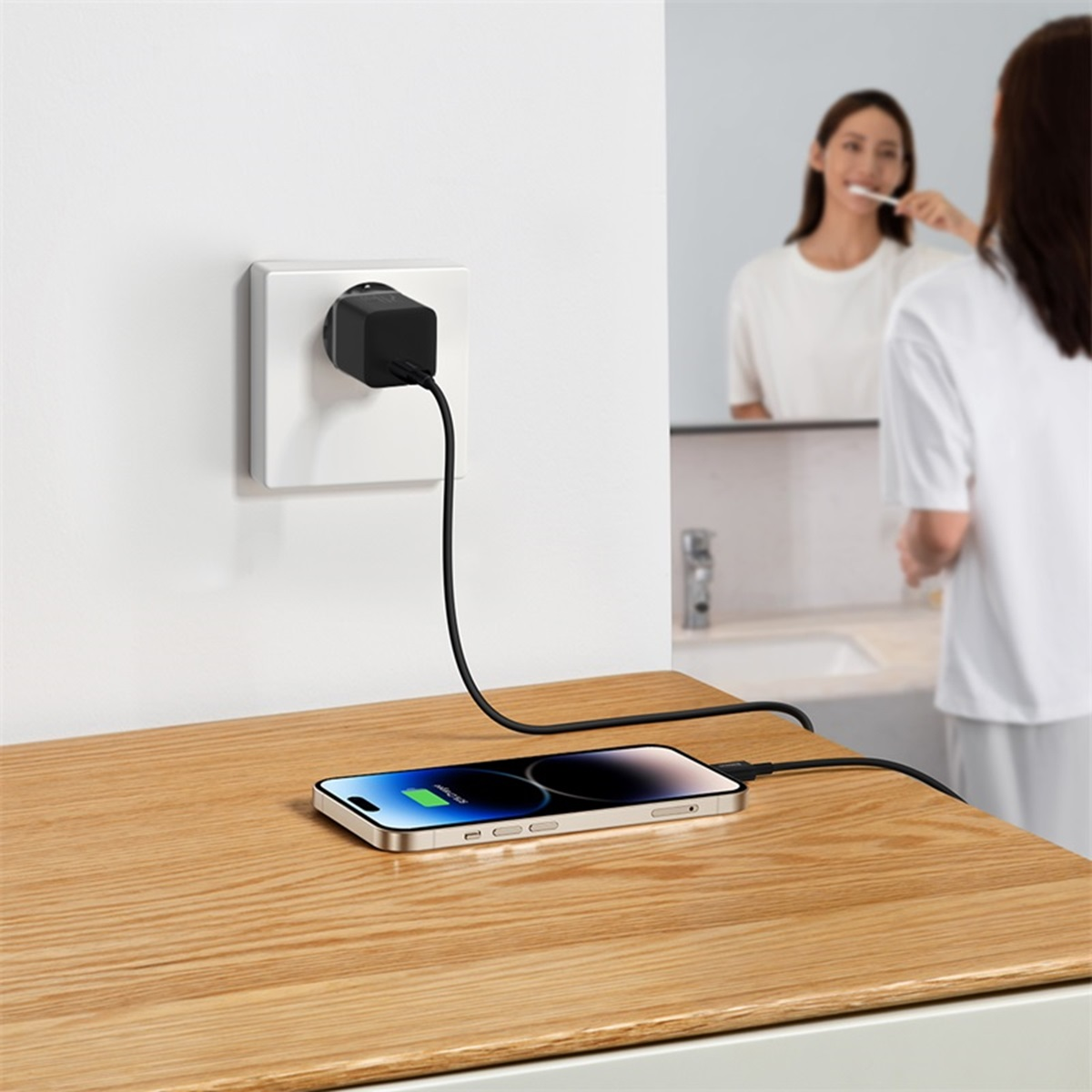 Teléfono cargando en mesa de madera. Conectado a toma de corriente. Mujer cepillándose dientes detrás.