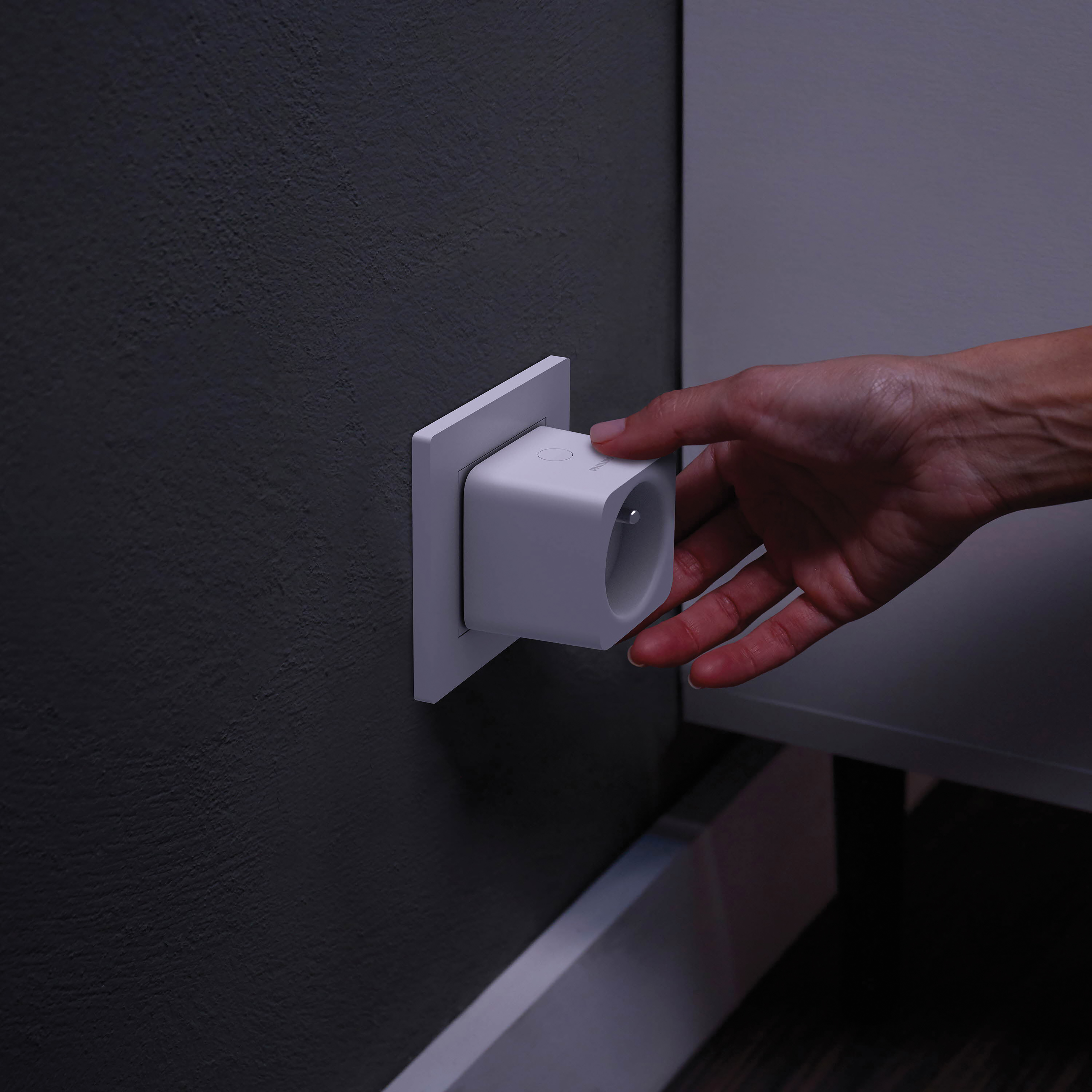 Una mano inserisce una presa intelligente bianca in una presa a muro grigio scuro. Cornice bianca visibile.
