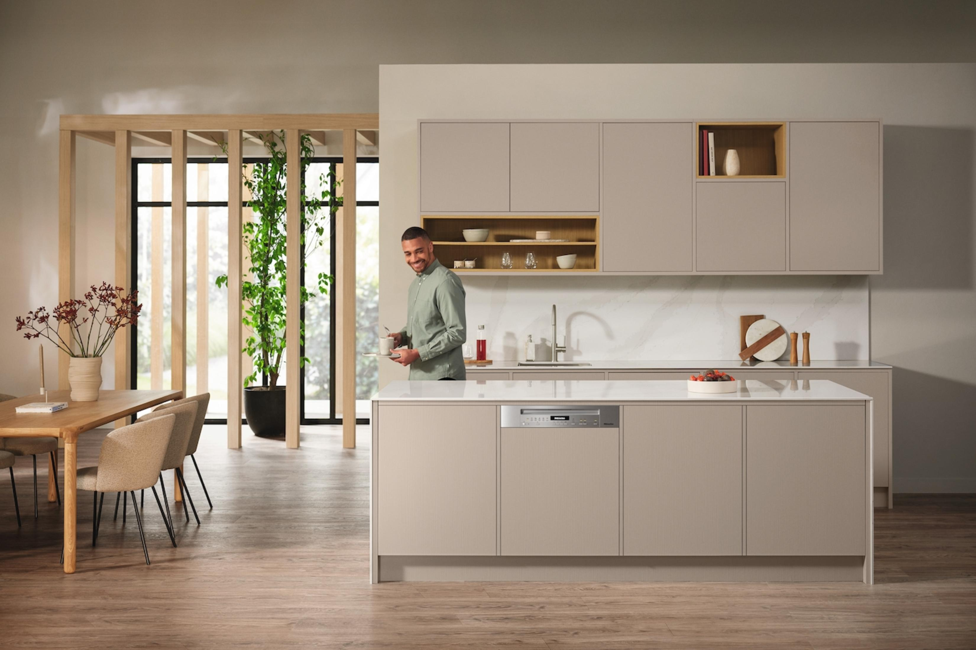 Eine moderne Küche mit einem lächelnden Mann. Beige Schränke, eine Insel und ein Holzrahmen sind sichtbar.