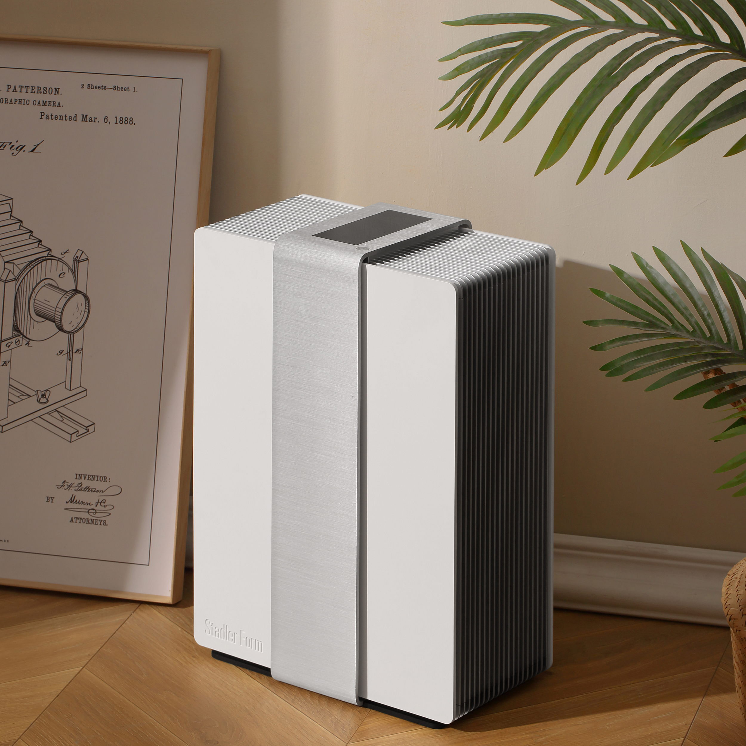 Air purifier with a silver band, next to a framed print and plant.