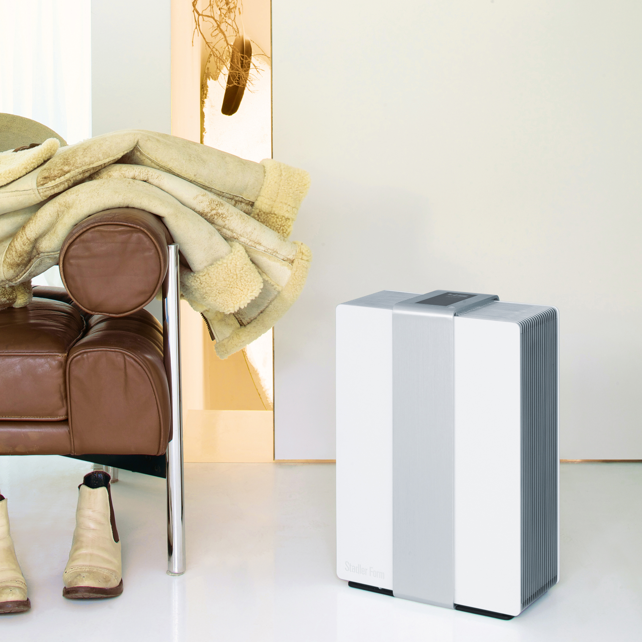 A white air purifier next to a brown leather sofa and a coat. It is on a white floor.