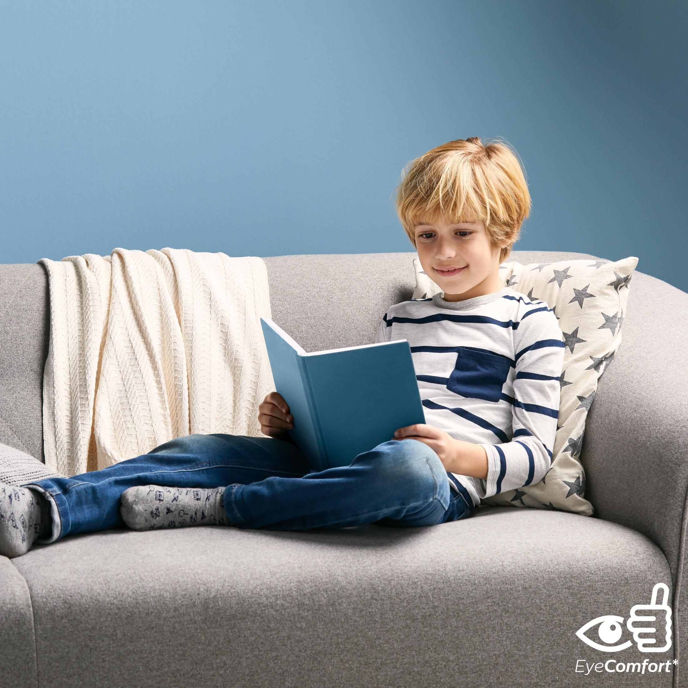 Een jongen leest een boek op een grijze bank met een kussen, met een blauwe muur op de achtergrond.