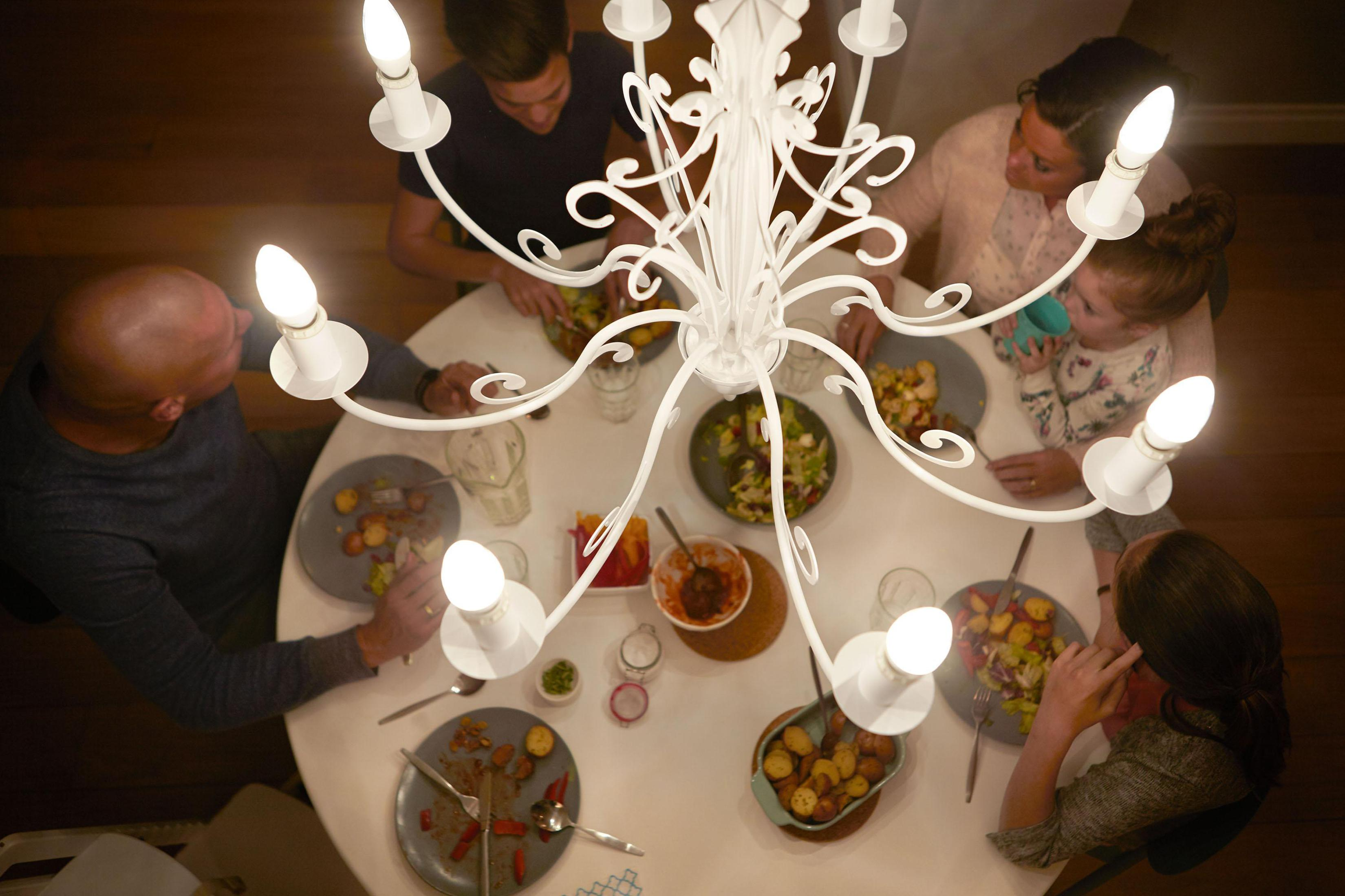 Familie dineert aan een witte tafel onder een kroonluchter. Borden met eten zichtbaar. Overhead shot.