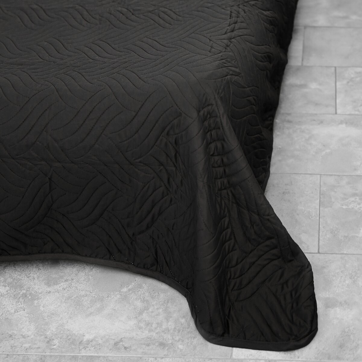 Eine schwarze Steppdecke bedeckt ein Bett. Die Steppdecke hat ein wellenförmiges Muster. Auf grauen Fliesen.