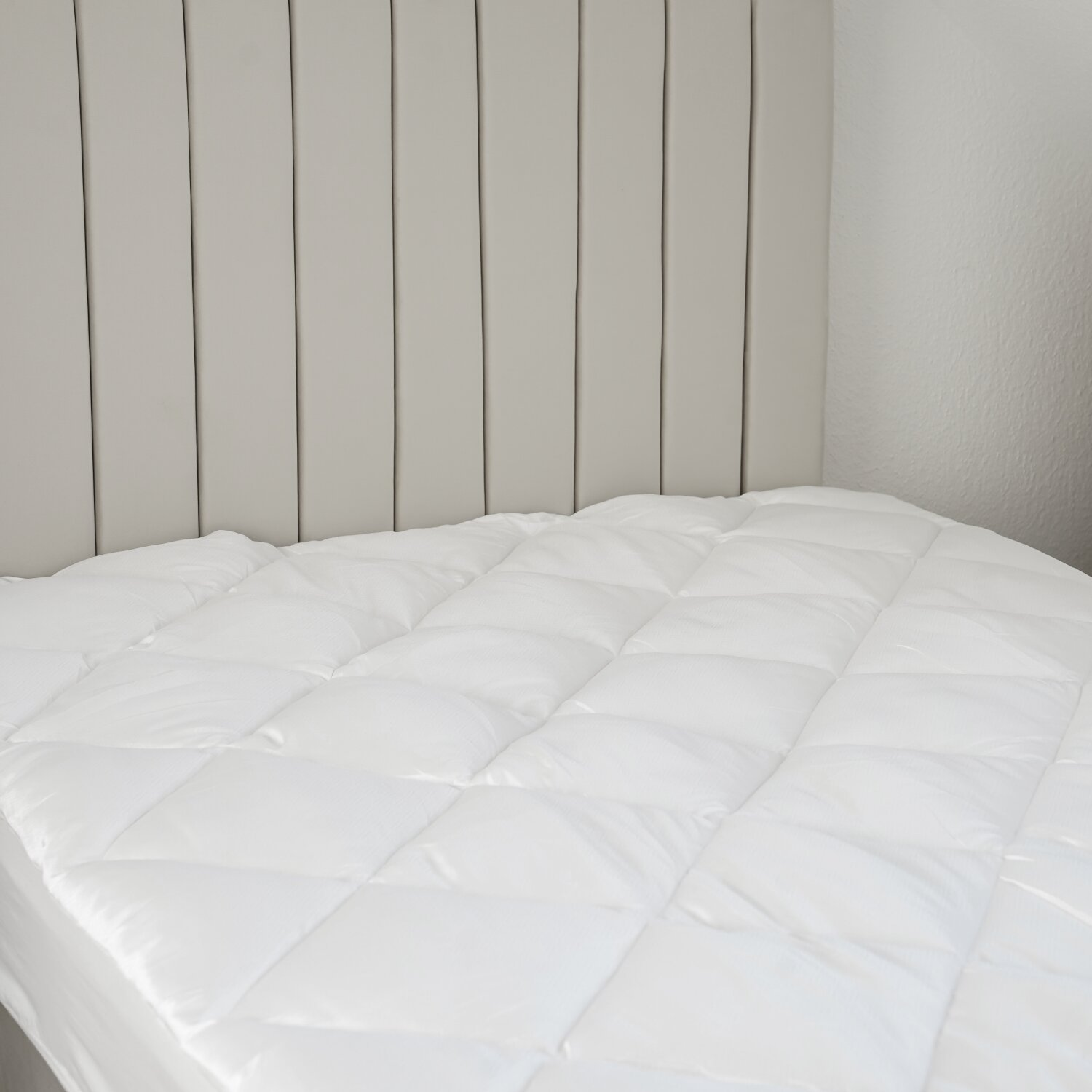 Weiß gesteppte Matratze auf einem Bett. Vertikales, hellgraues Kopfende dahinter. Weiße Wände.