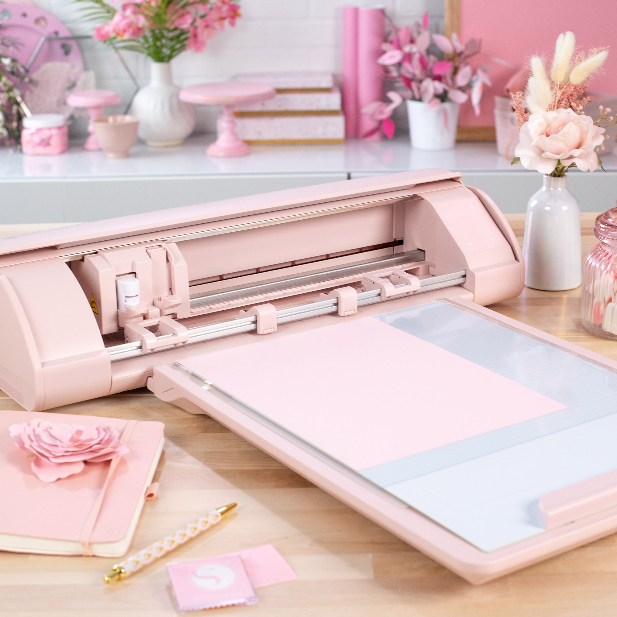Eine rosa Bastelmaschine mit Papier, Stiften und Blumen auf einem Holztisch.