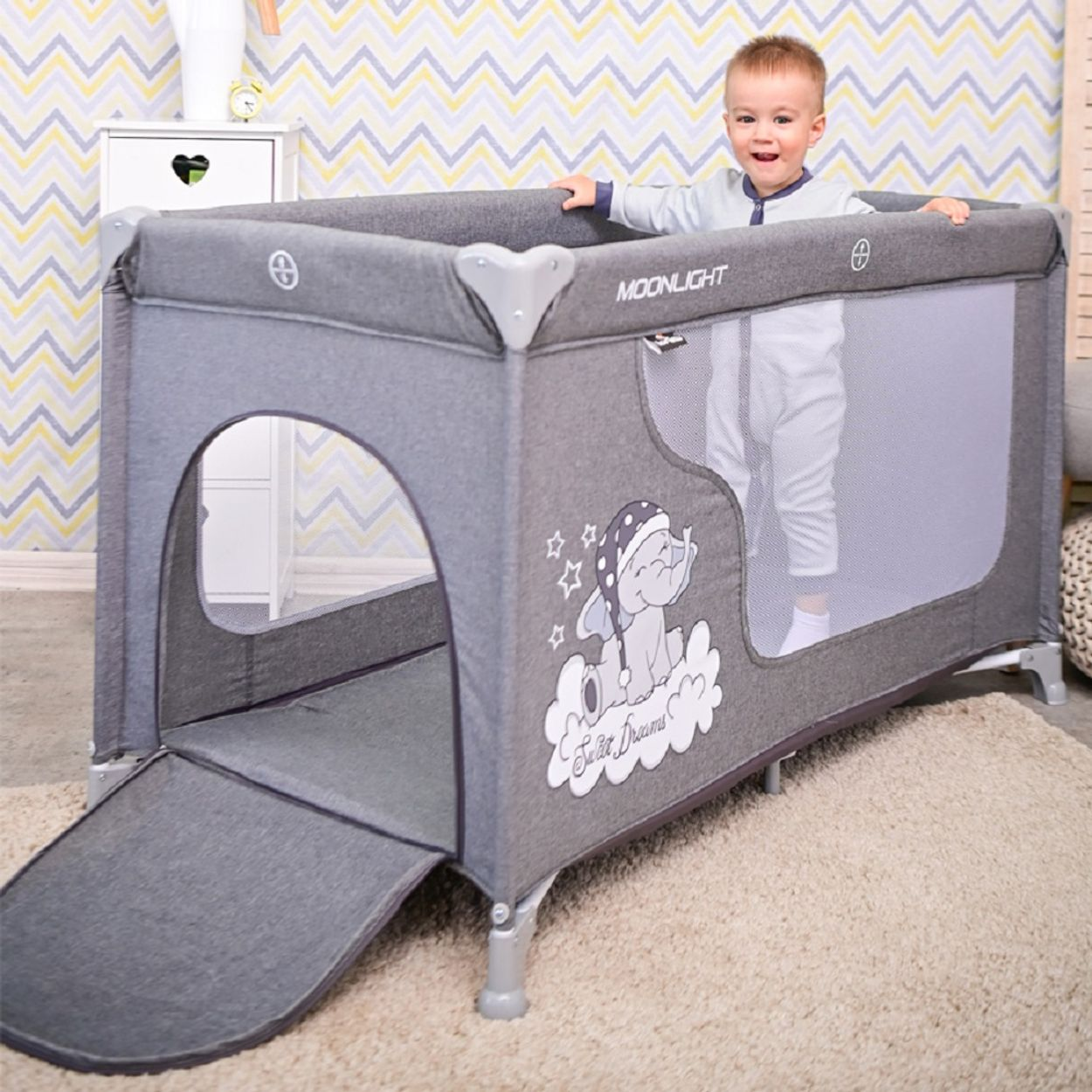 Grauer Baby-Reisebett mit Kind, das darin steht und lächelt. Das Bett ist offen.