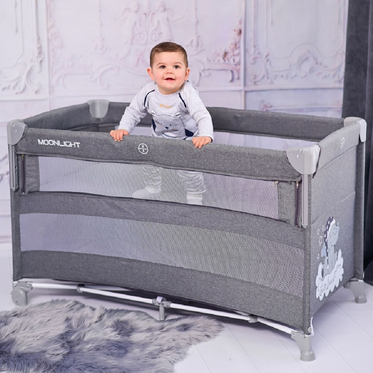 Ein Baby steht in einem grauen Kinderbett. Das Bett hat eine Gitterseite und einen grauen Fellteppich.