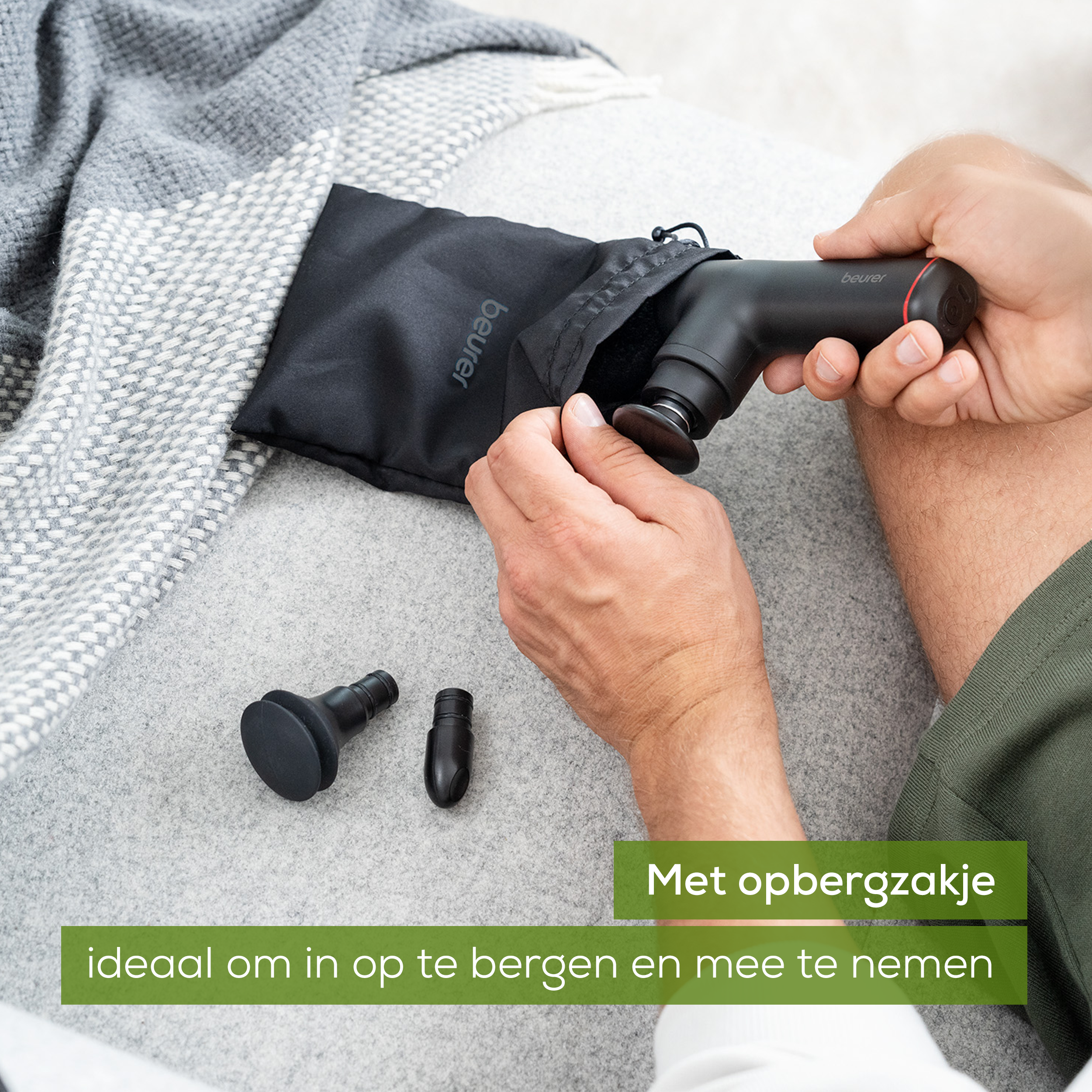 Mani che tengono una pistola per massaggio nera, con accessori. Divano e coperta grigi.