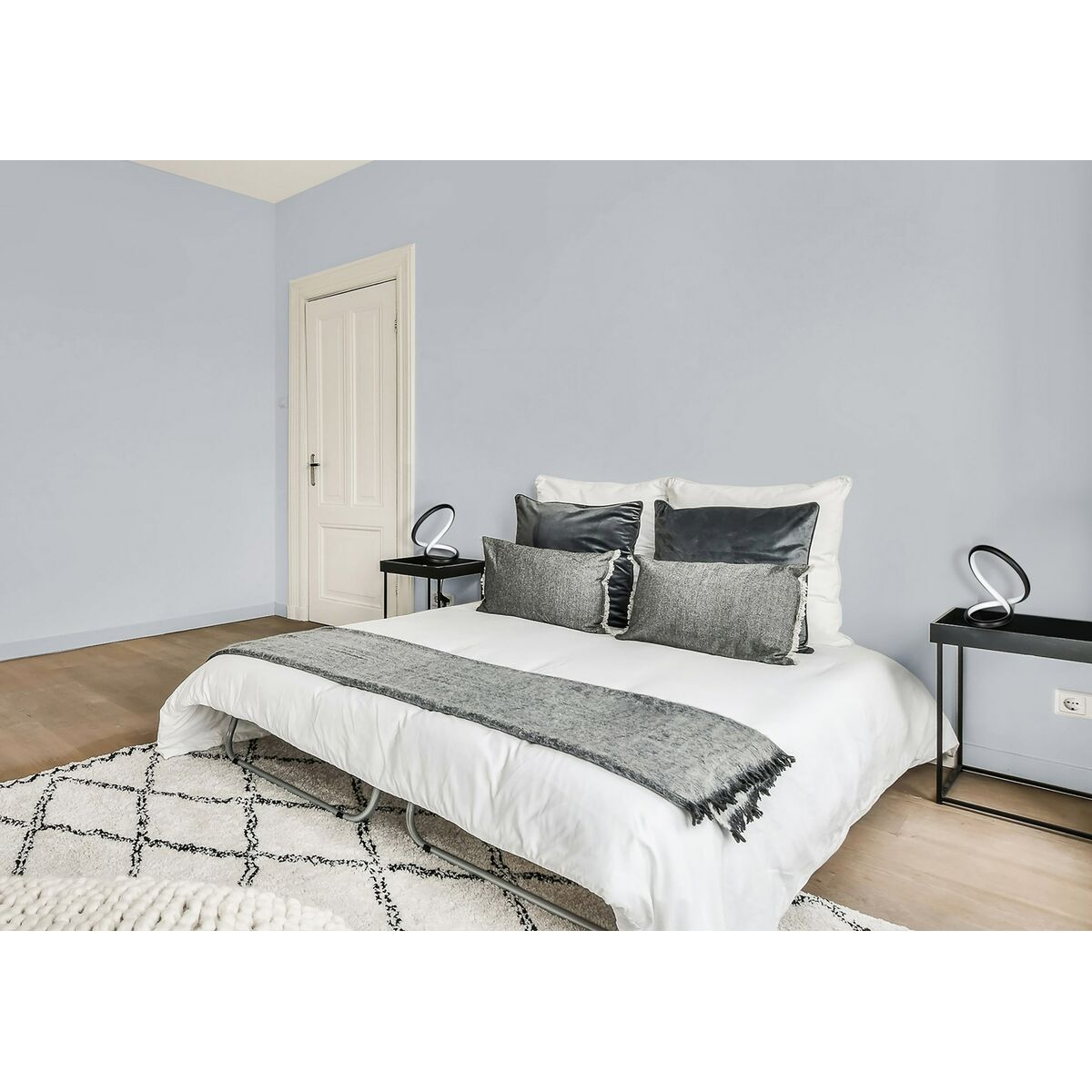 Dormitorio con cama, dos mesitas de noche y una alfombra. Las paredes son de color azul claro. Hay una puerta.