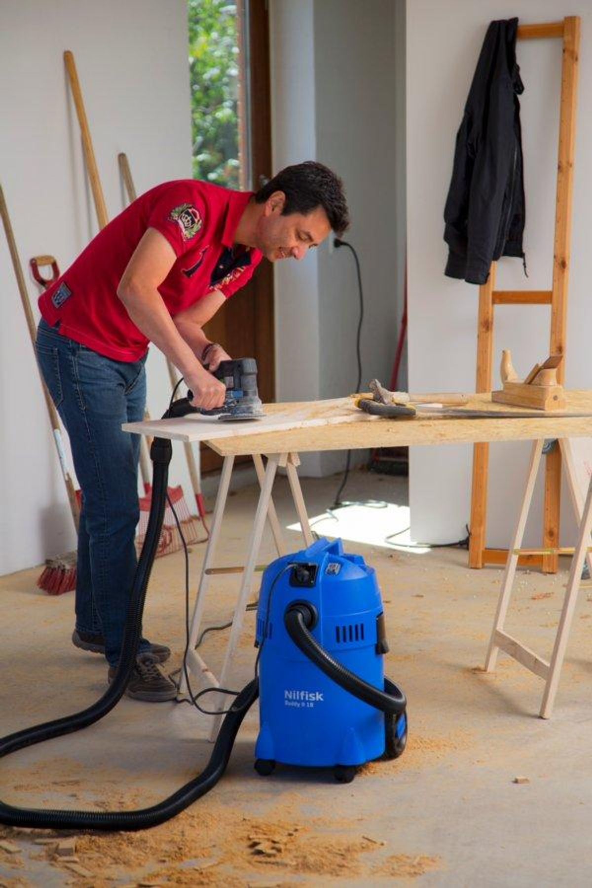 Hombre lijando madera sobre mesa con aspiradora cerca. Herramientas y chaqueta en la habitación.