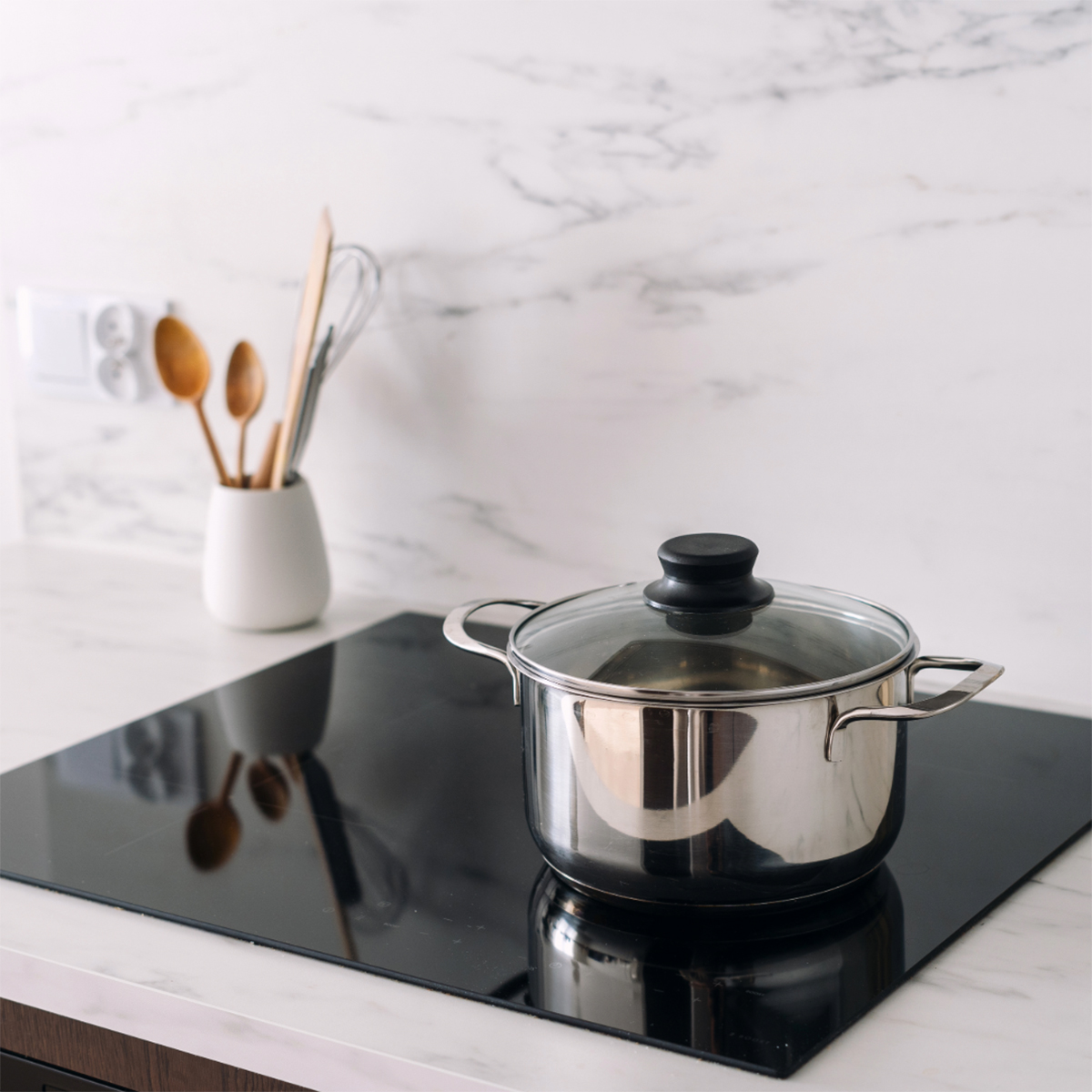 Olla de acero inoxidable con tapa de vidrio sobre vitrocerámica negra, utensilios de madera.